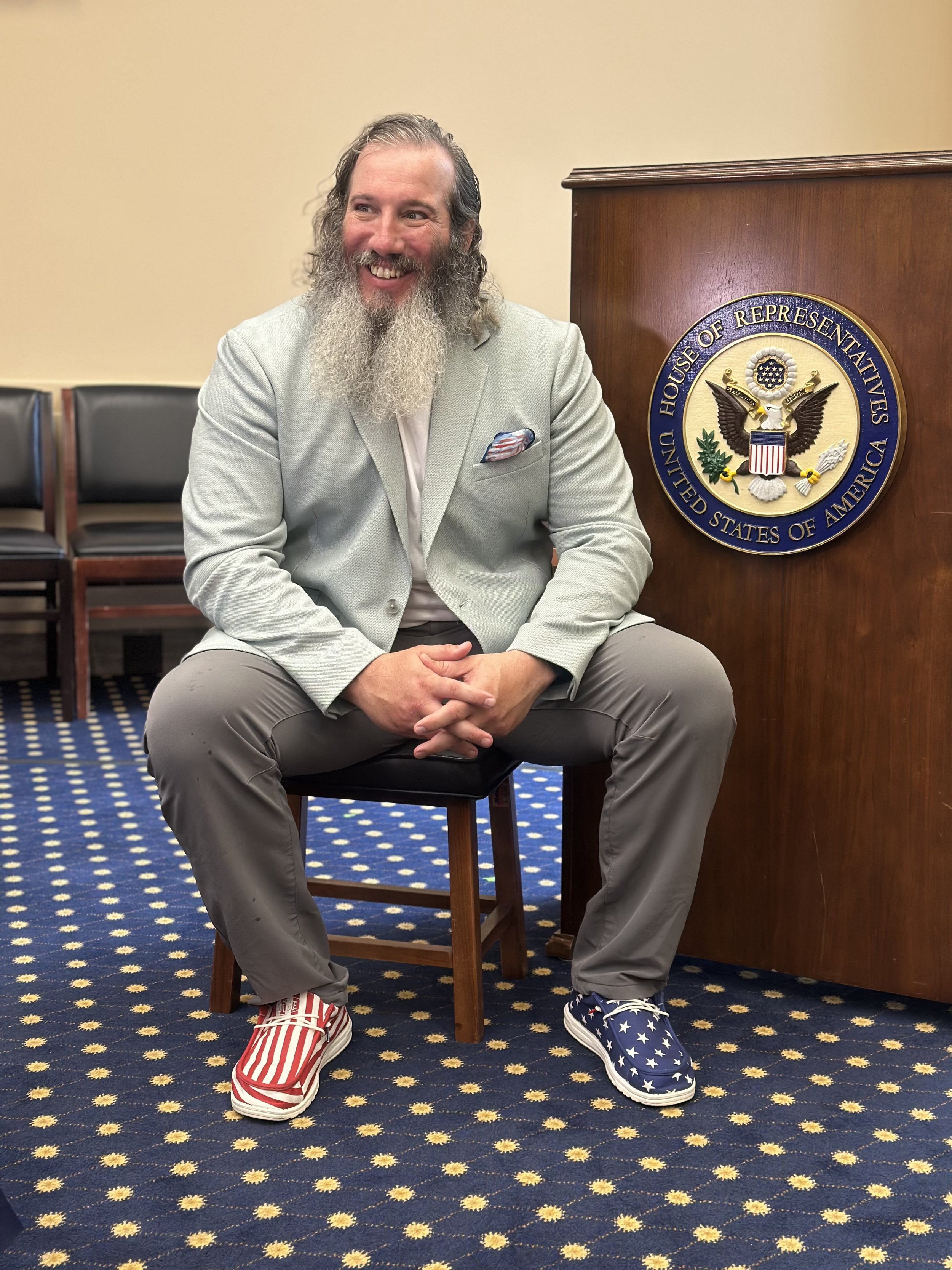 A man with a long beard and long hair, sitting on a chair in a room with a blue carpet with yellow flower patterns. He is wearing a light gray blazer, beige pants, and American flag-themed sneakers. He is smiling, with a handshake pose, and there is 