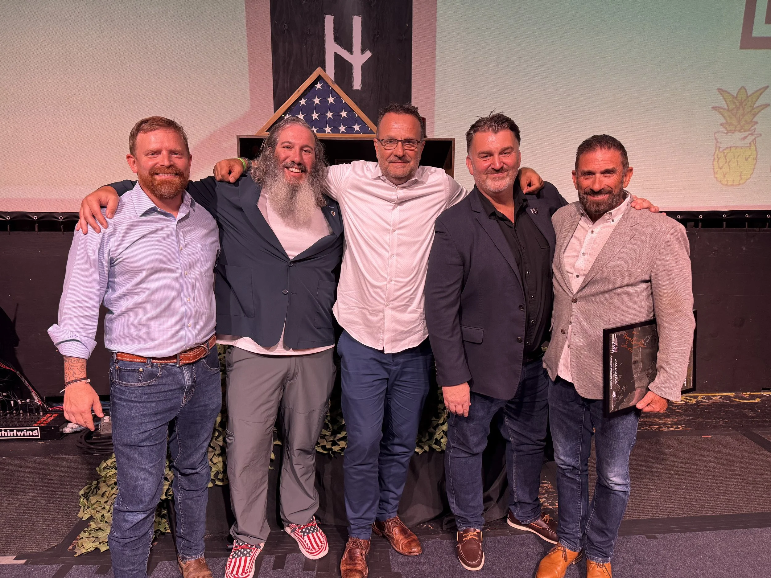 Five men standing together on stage with their arms around each other, smiling. Behind them is a fitment with an American flag design, a pineapple logo, and the letters "LY". One man on the far right holds a book.