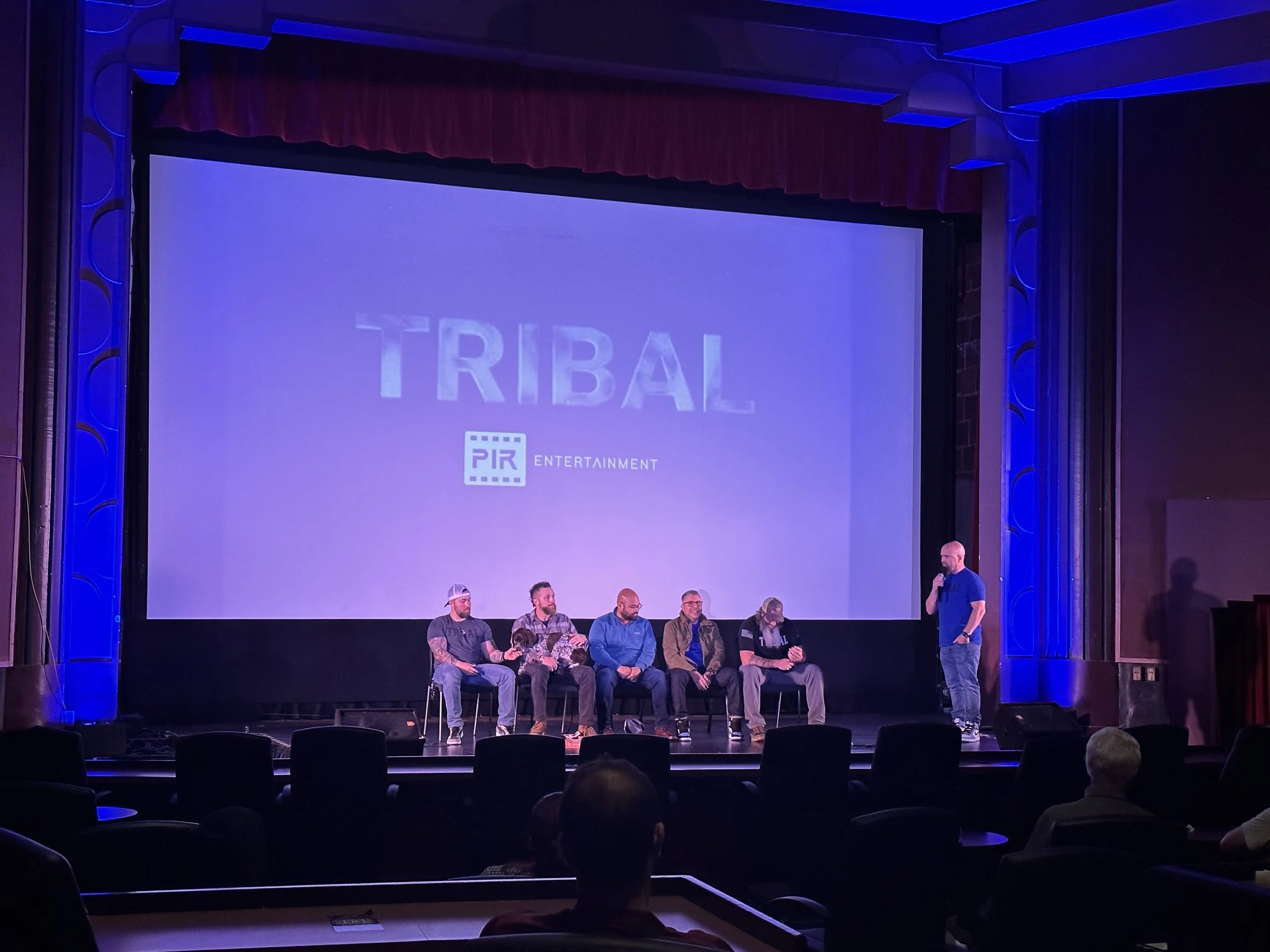 A stage with a large screen displaying the word 'TRIBAL' and 'PIR ENTERTAINMENT' logo. There are six people sitting on chairs on stage and one person standing and speaking into a microphone. The stage is lit with blue lighting and has a red curtain a