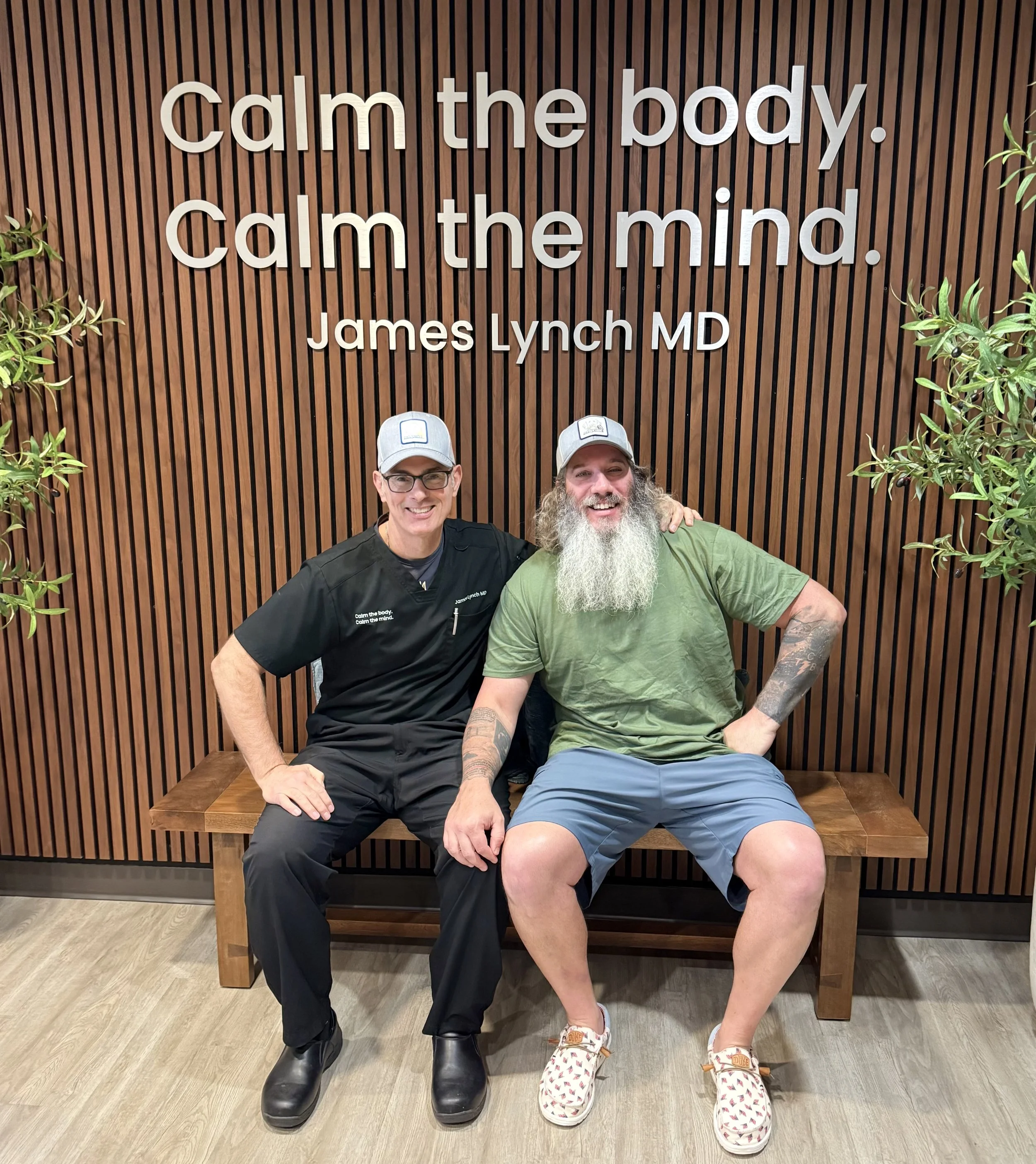 Two men sitting on a wooden bench in front of a wooden slat wall. The man on the left is wearing a black medical scrubs and black boots, and the man on the right has a long beard, tattoos, a green T-shirt, shorts, and sneakers with animal print. The 