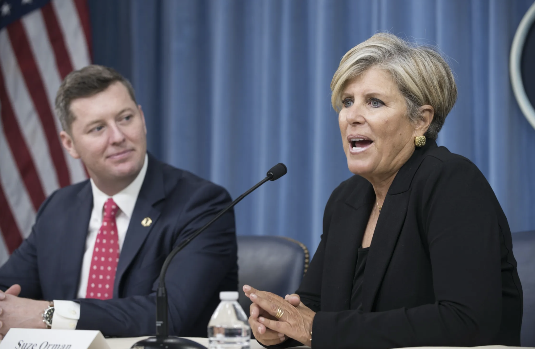 Two people sitting at a panel discussion with microphones. The woman on the right is speaking, wearing a black blazer and gold earrings. The man on the left is listening, dressed in a navy suit and red polka dot tie. An American flag is visible in the background.