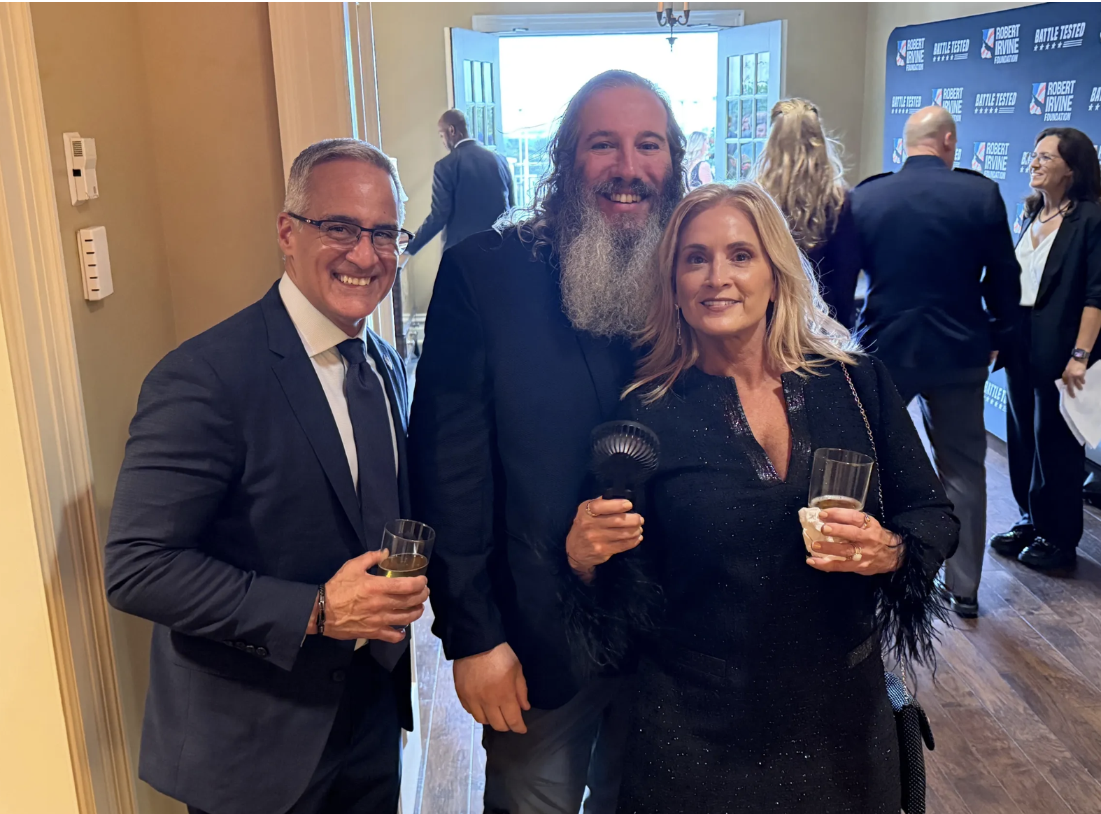 Three people at a formal event, a man in a suit with glasses holding a drink on the left, a woman with blonde hair holding a microphone and a drink on the right, and a man with a long beard in the middle, smiling. In the background, people are gather