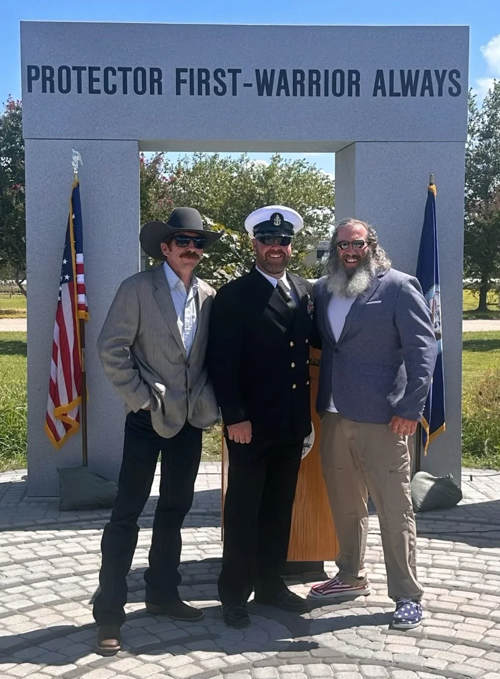 Three men in suits and uniforms standing under a memorial arch with the inscription "PROTECTOR FIRST-WARRIOR ALWAYS." Flags on either side of the arch.