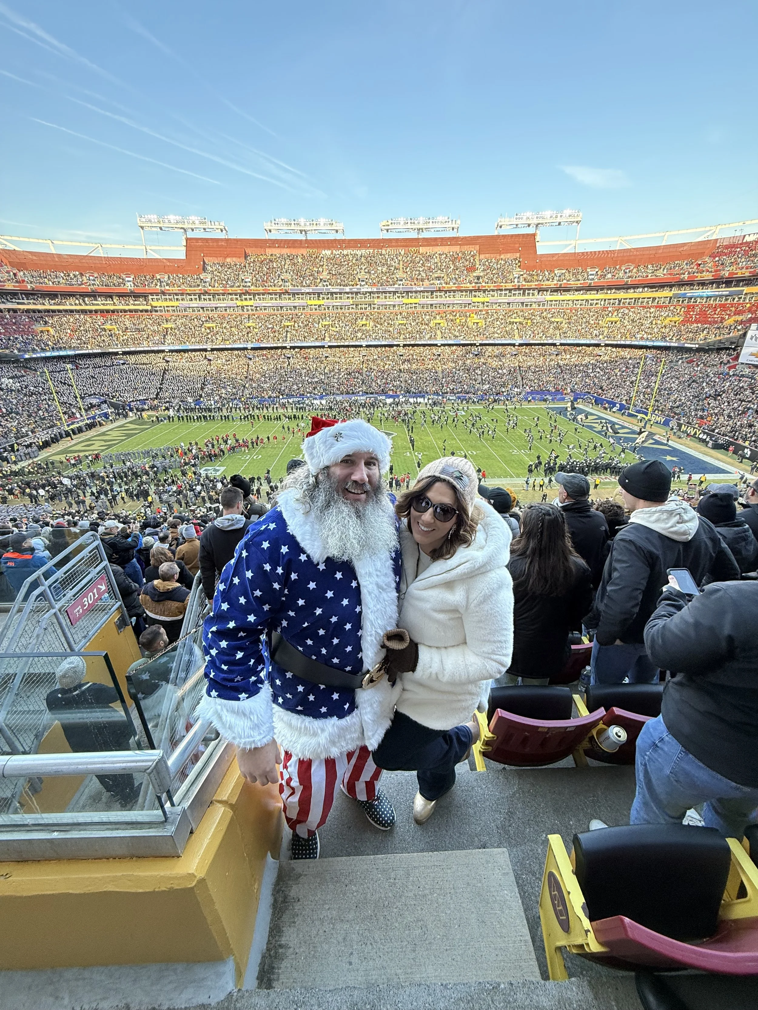 People enjoying a football game at a stadium, with two individuals dressed in holiday costumes, one in a Santa Claus outfit and the other in a white coat, posing for a photo at a stadium entrance or balcony with a football field visible in the backgr