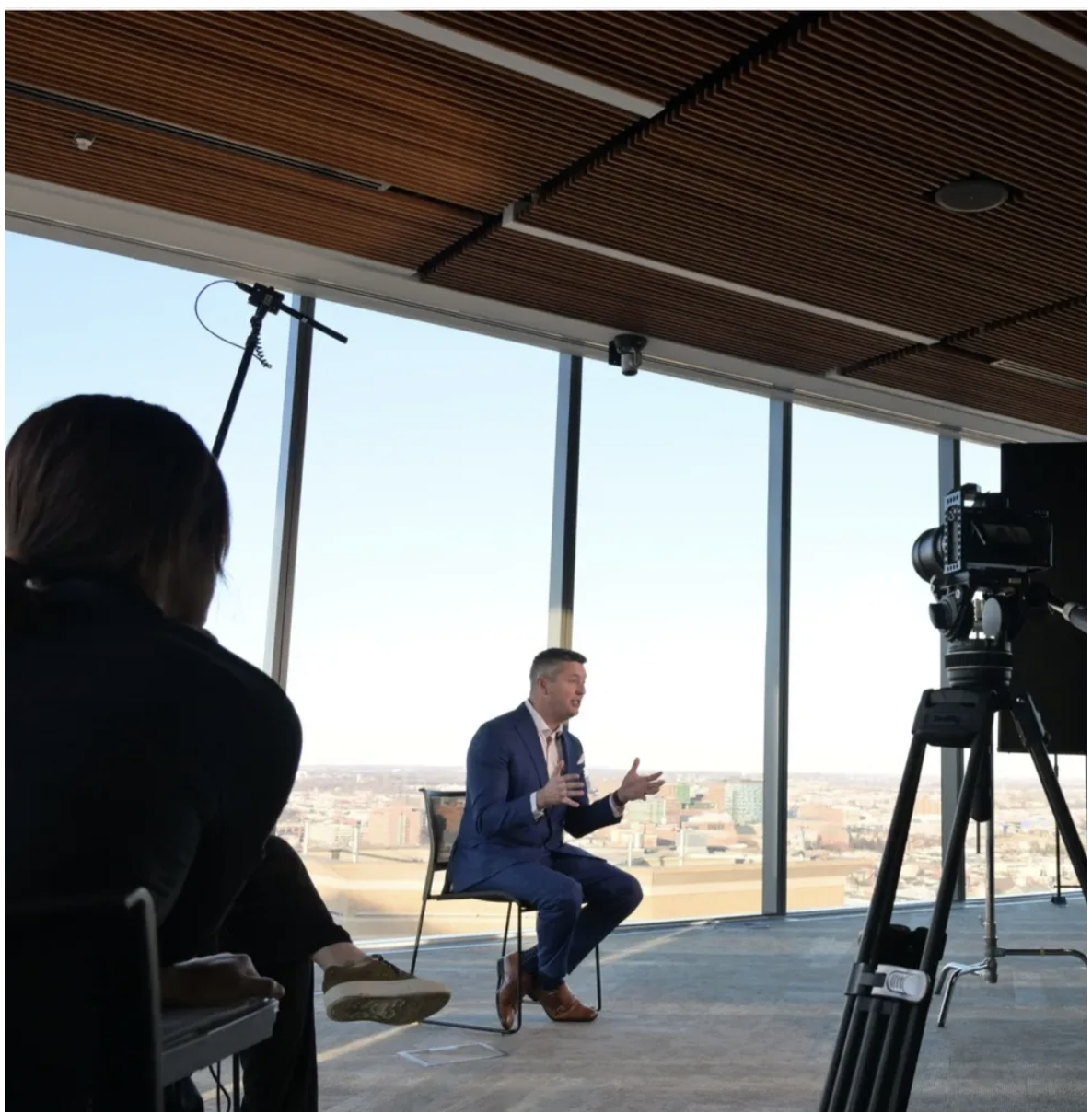 A man in a blue suit is giving a presentation or interview in a high-rise building with floor-to-ceiling windows showing a cityscape. A woman and another person are seated nearby, and a camera is recording the scene.