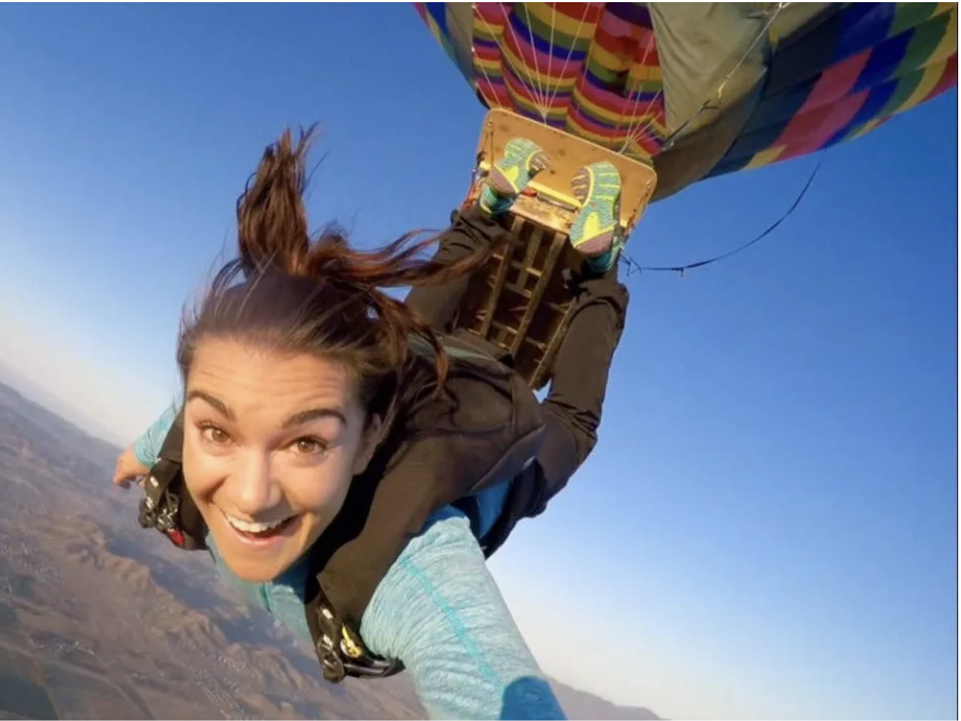 A woman with long brown hair skydiving, smiling, wearing a black jumpsuit, with a colorful parachute above her in the clear blue sky.