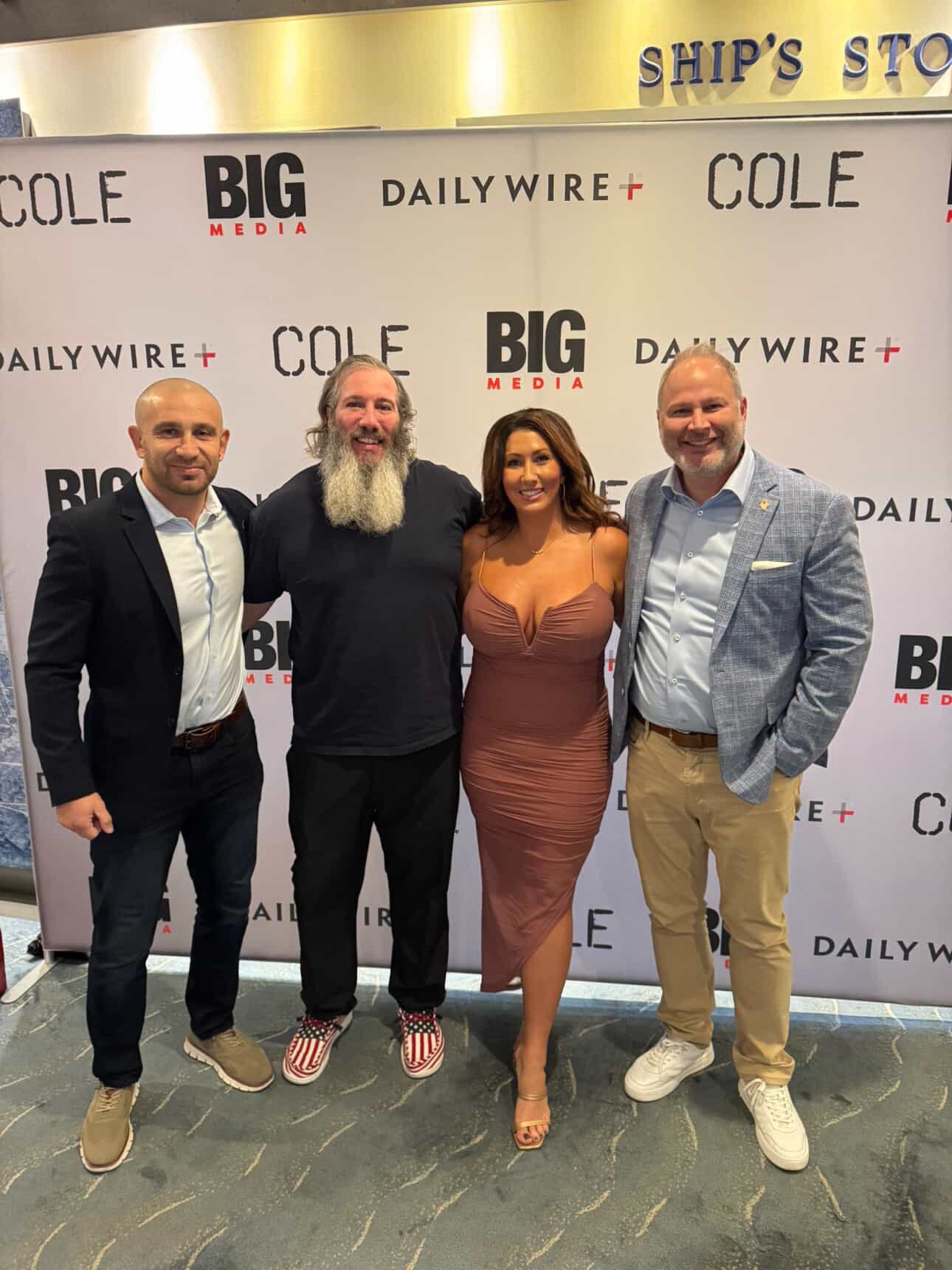 Four people posing together in front of a backdrop with media logos at an event, with three men and one woman all smiling and dressed in business or cocktail attire.
