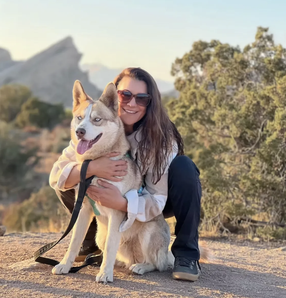 A woman in sunglasses and a beige hoodie kneels beside a Siberian Husky with blue eyes on a mountain trail during sunset.