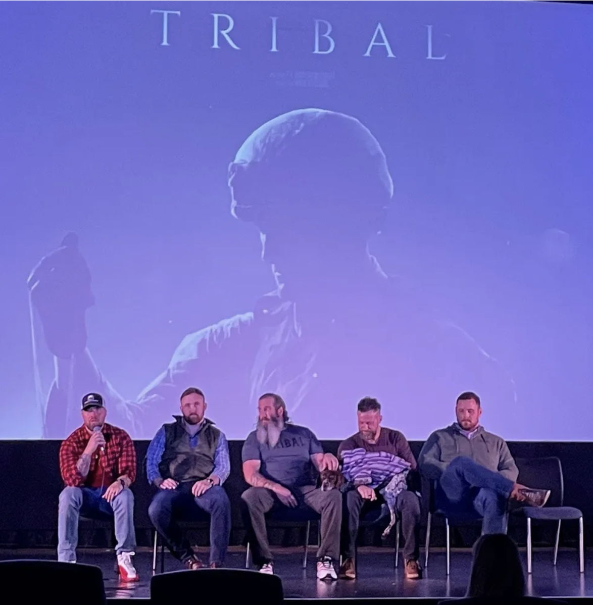 Five men sitting on a stage in front of a large screen displaying the word 'TRIBAL' and an image of a person holding an axe. One man is speaking into a microphone, and a dog is sitting next to one of the men.
