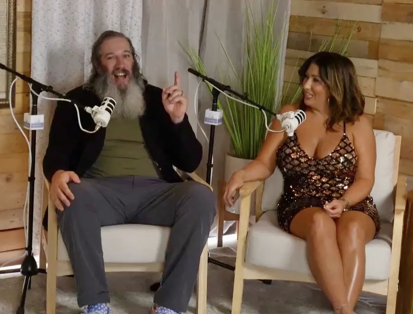Two people, a man with a long gray beard and a woman with shoulder-length brown hair, are sitting in chairs in front of microphones, engaging in conversation. The man is wearing a dark blazer, green shirt, and gray pants, and is gesturing with his ri