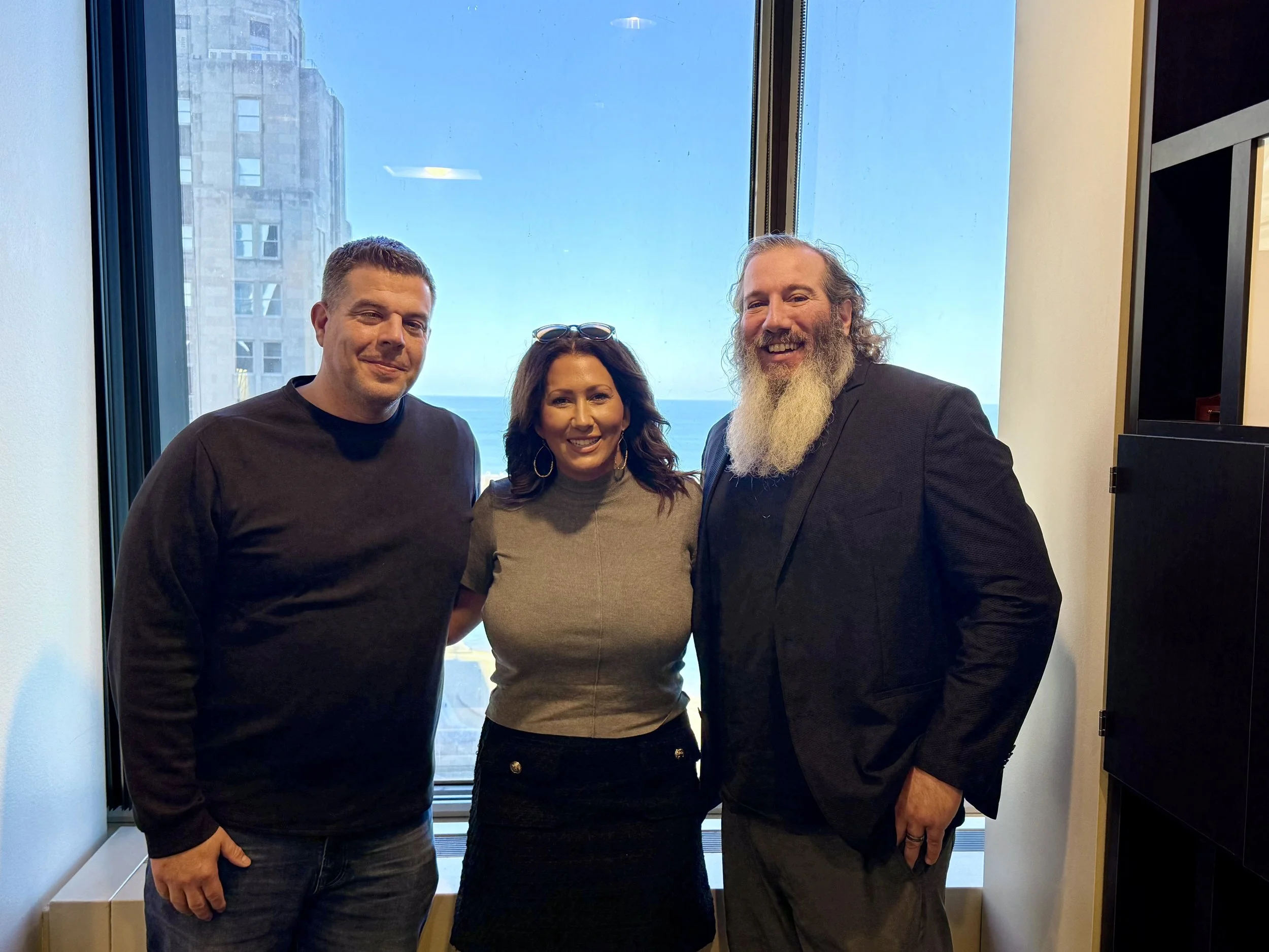 Three smiling people, two men and a woman, standing together indoors near a large window with a cityscape view in the background.