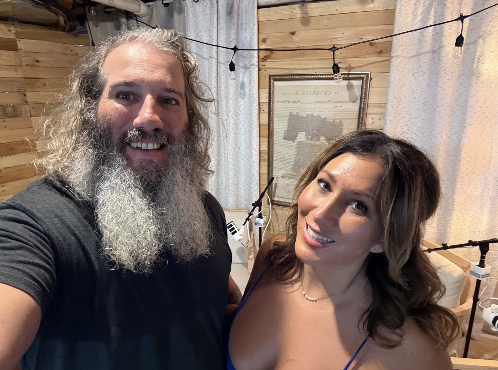 A man with long curly gray hair and beard taking a selfie with a woman with long wavy brown hair in a room with wooden walls and string lights.