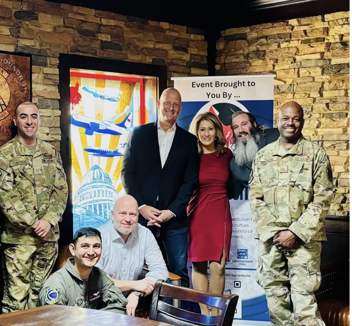 Group of seven people, including two in military uniforms, standing and sitting in a room with brick walls and a stained glass window, gathered for a photo at an event.