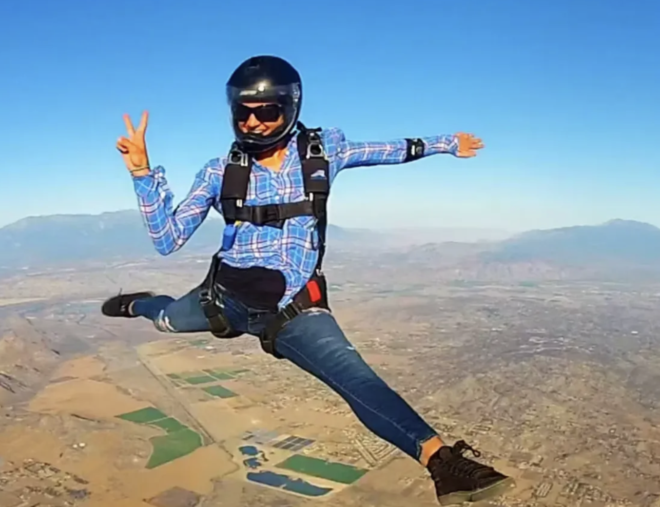 Person wearing a helmet, sunglasses, and a plaid shirt skydiving with arms and legs spread out over a landscape of fields and mountains.