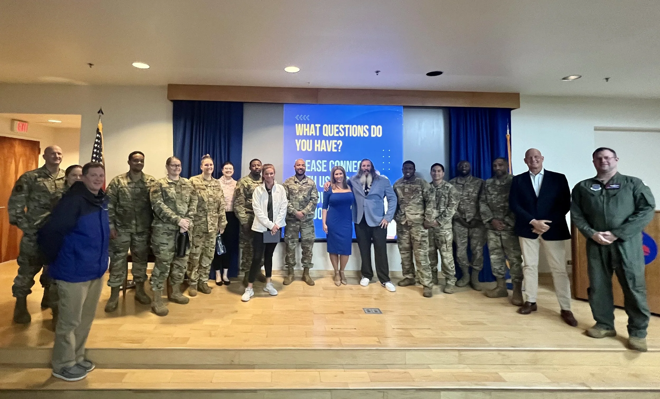 Group of people, including military personnel and civilians, standing on a stage in front of a large screen with the text "What questions do you have?" and "Please connect with us."