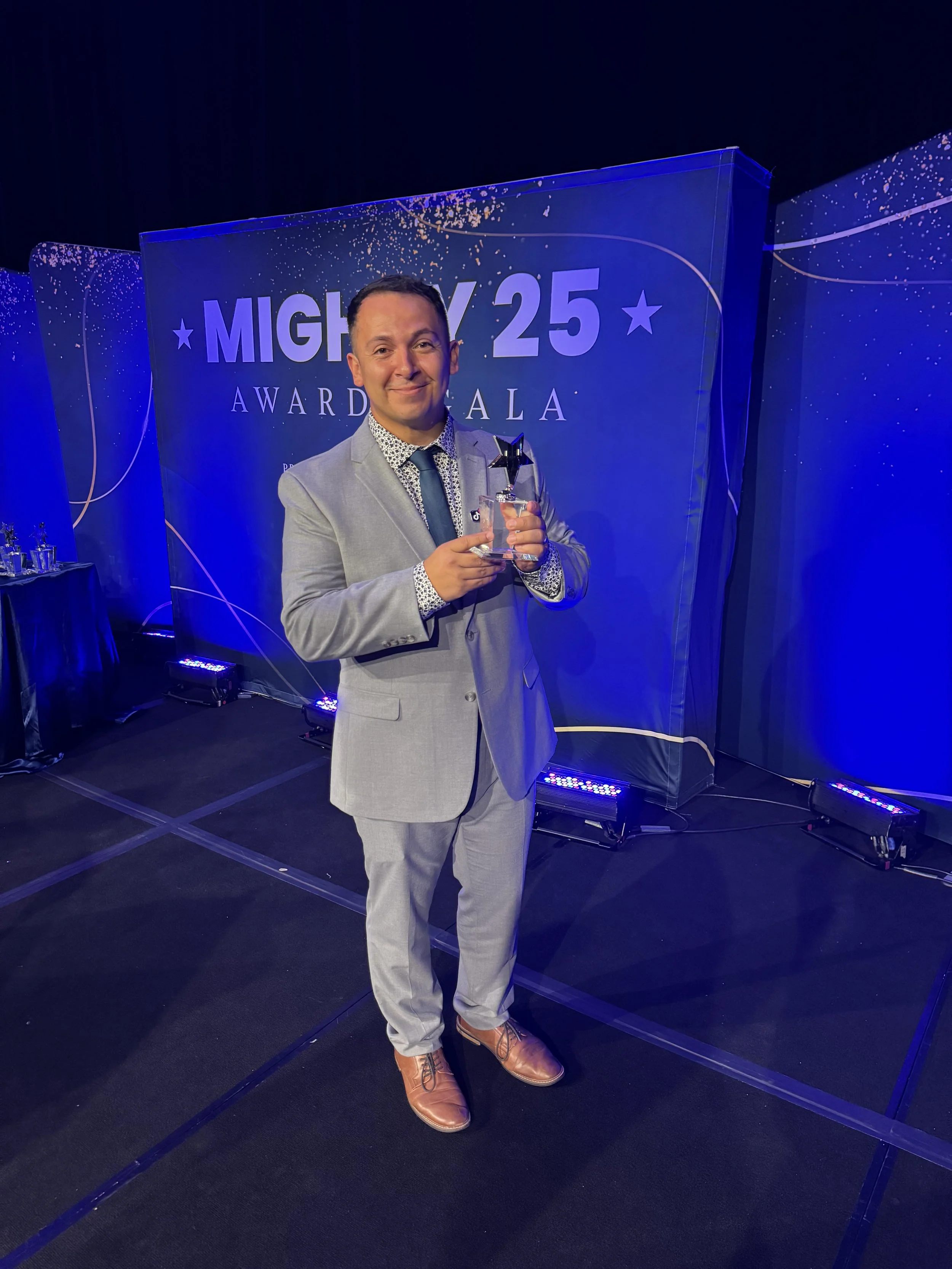 A man in a light gray suit and brown boots holding an award with a star on top, standing in front of a blue backdrop with the words 'MIGHTY 25 AWARD GALA' and decorative white lines and dots.