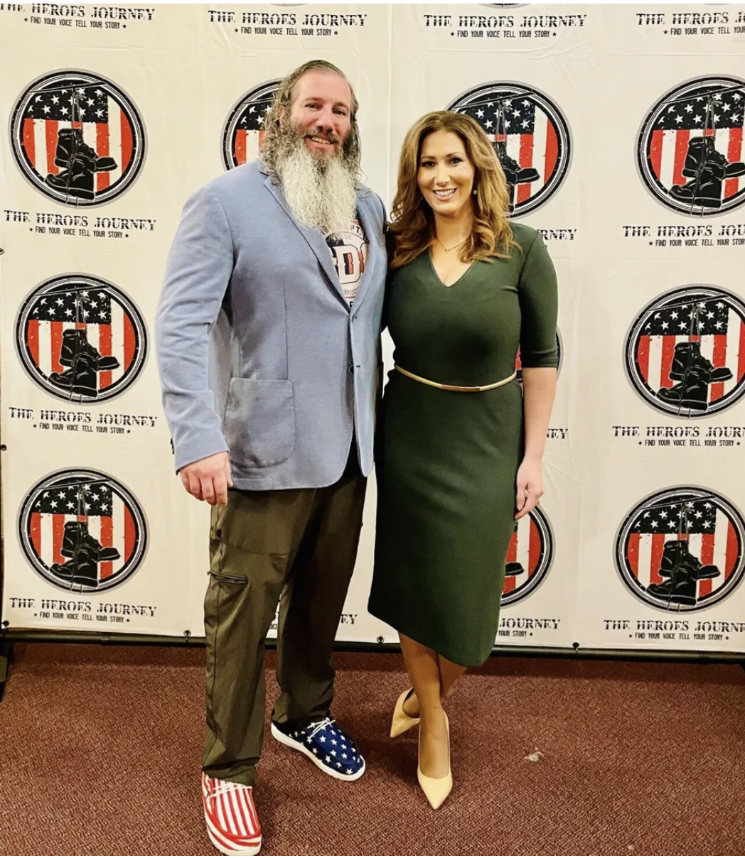 A man with long gray beard and a woman are standing together in front of a backdrop with a logo that features a black boot and an American flag with the words "The Heroes Journey" and "Find your voice tell your story." The man is wearing a blue blaze