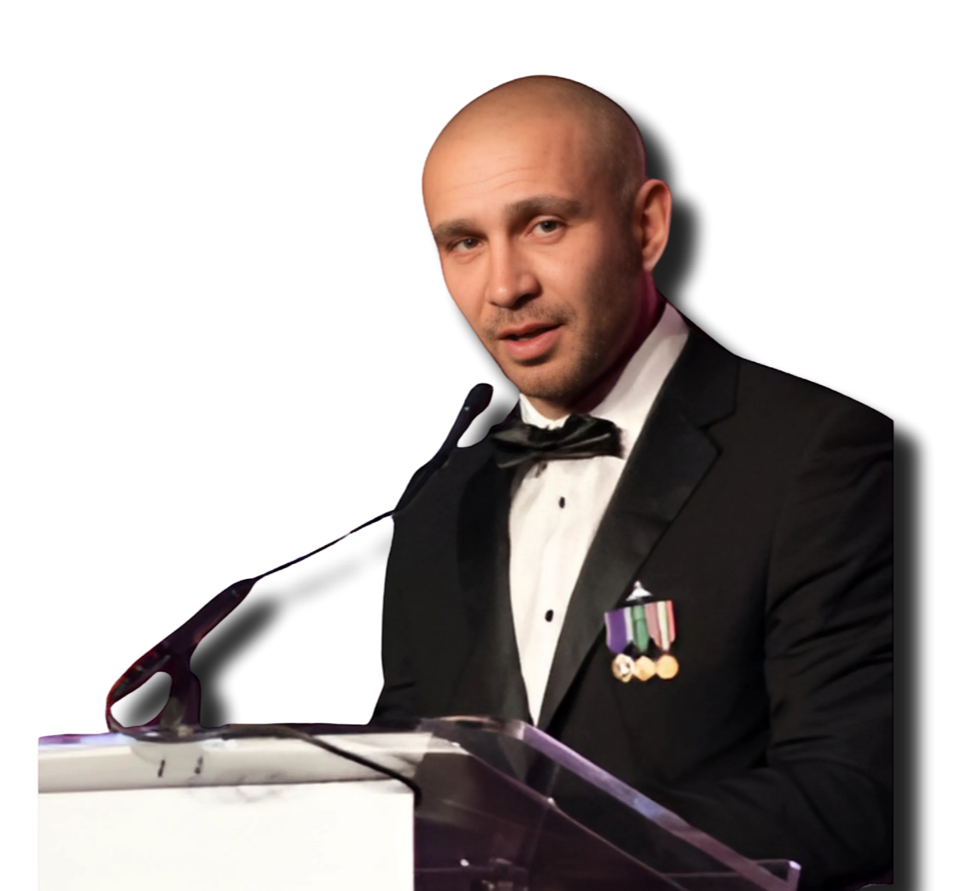 A man in a tuxedo with medals on his chest speaking at a podium with a microphone.