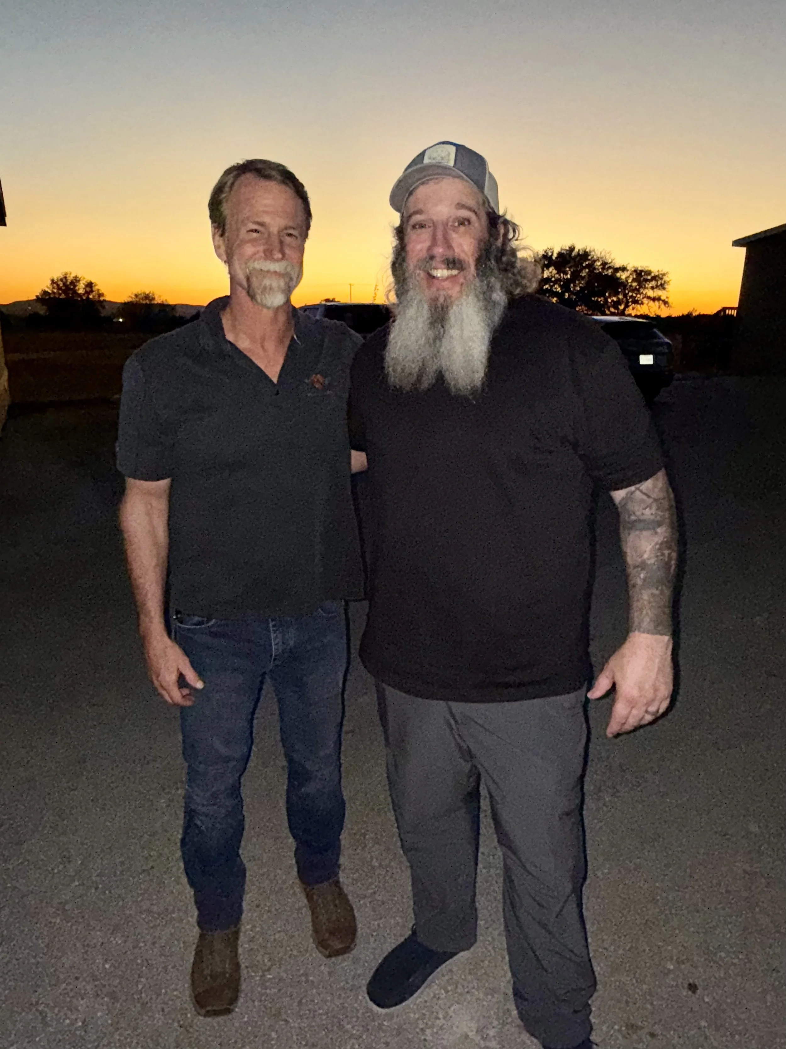 Two men standing outdoors at sunset, smiling at the camera. One has a beard and is wearing a hat and dark clothing; the other has a beard and is wearing a black shirt and jeans. There are trees and a car in the background.