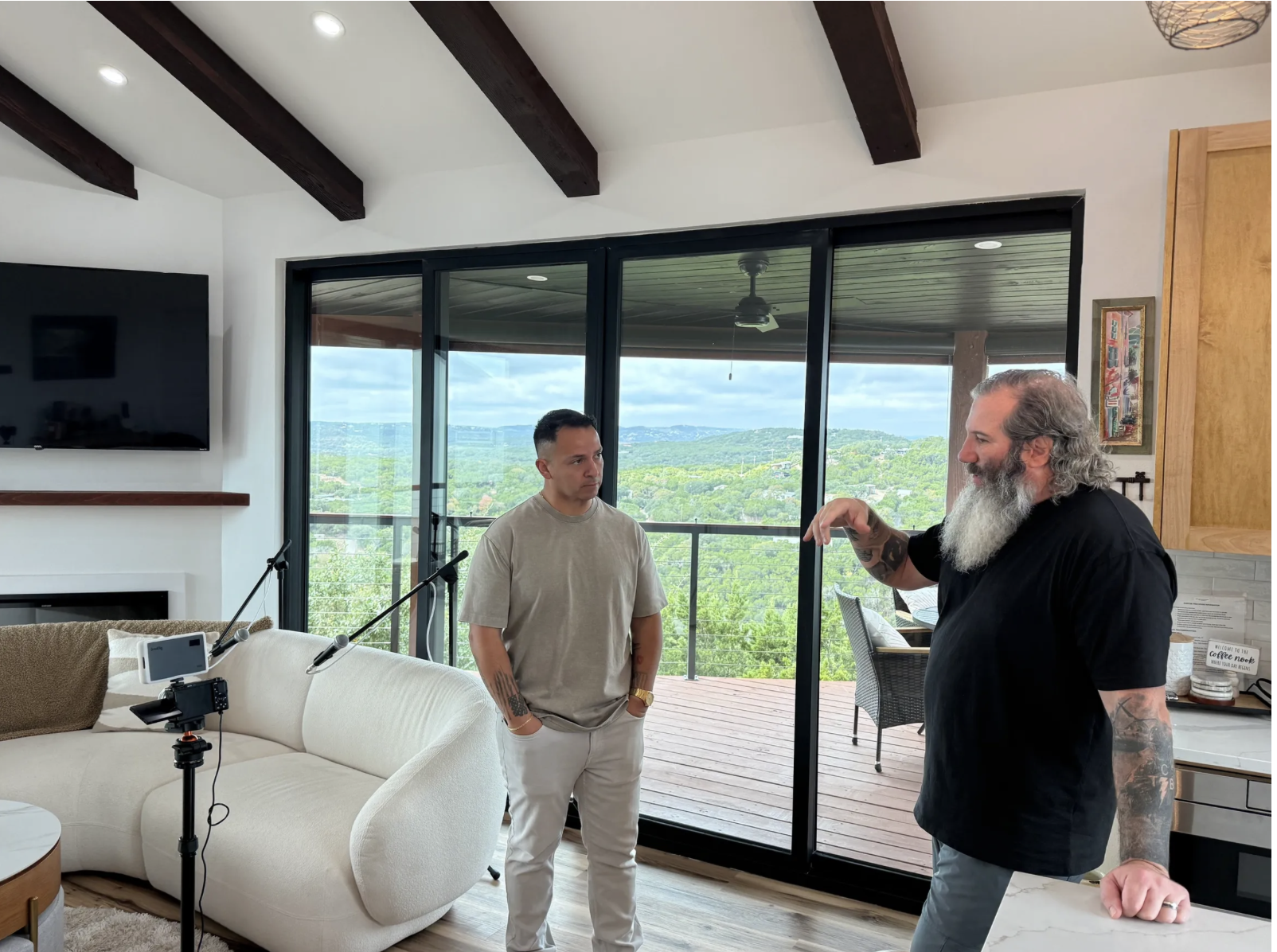 Two men are having a conversation in a bright living room with large sliding glass doors leading to a balcony overlooking a lush, green landscape.