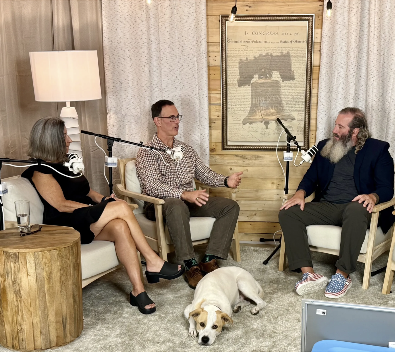 Three people engaged in a conversation in a cozy room, with a dog lying on the rug at their feet, and three microphones in front of them for recording.