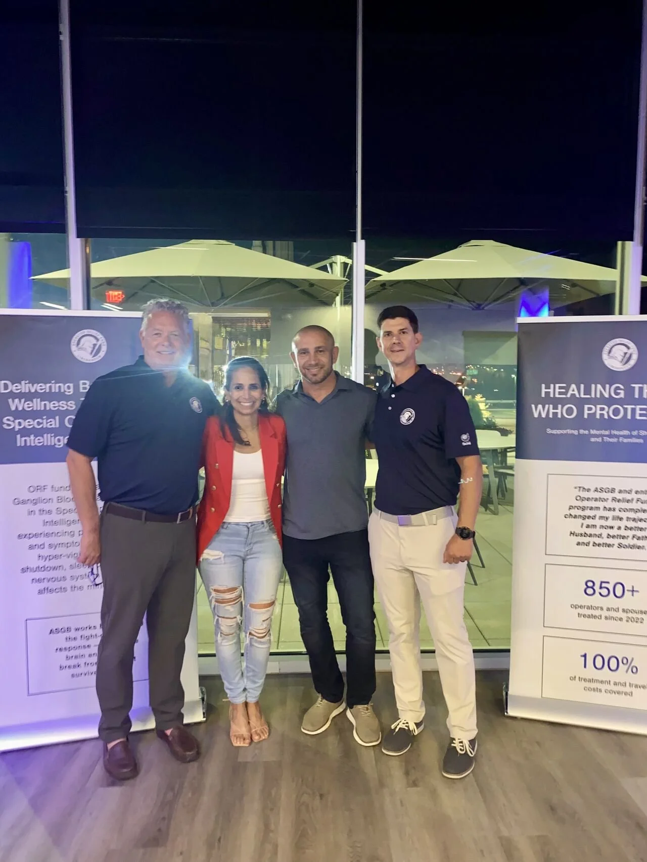 Four people standing together in front of informational banners at an event, with a glass window and umbrellas outside behind them.