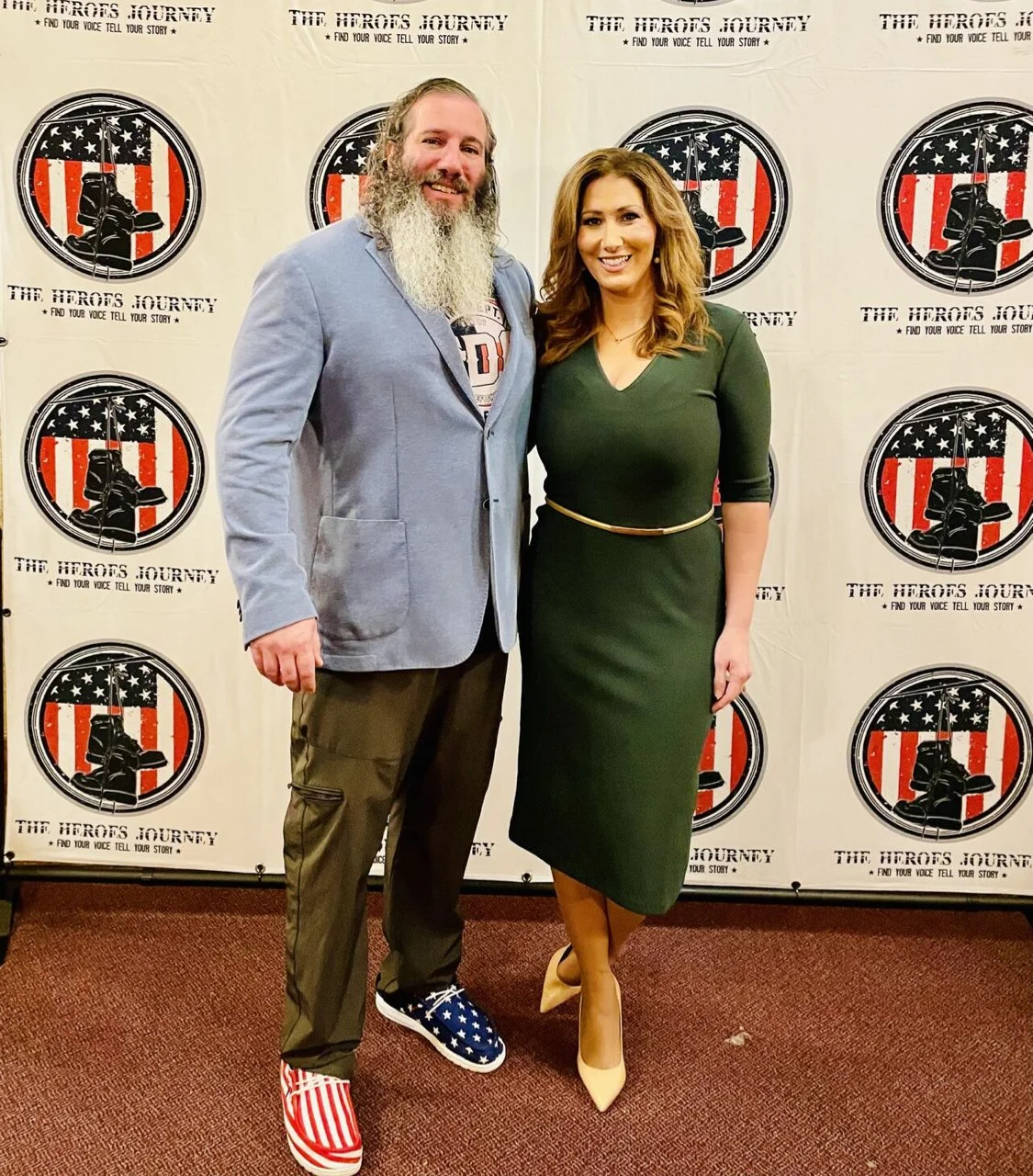 A man and woman stand together in front of a backdrop with a logo and the text "The Heroes Journey." The man has long gray hair and a beard, and he is wearing a gray blazer, brown pants, and patriotic shoes with the American flag. The woman has shoul