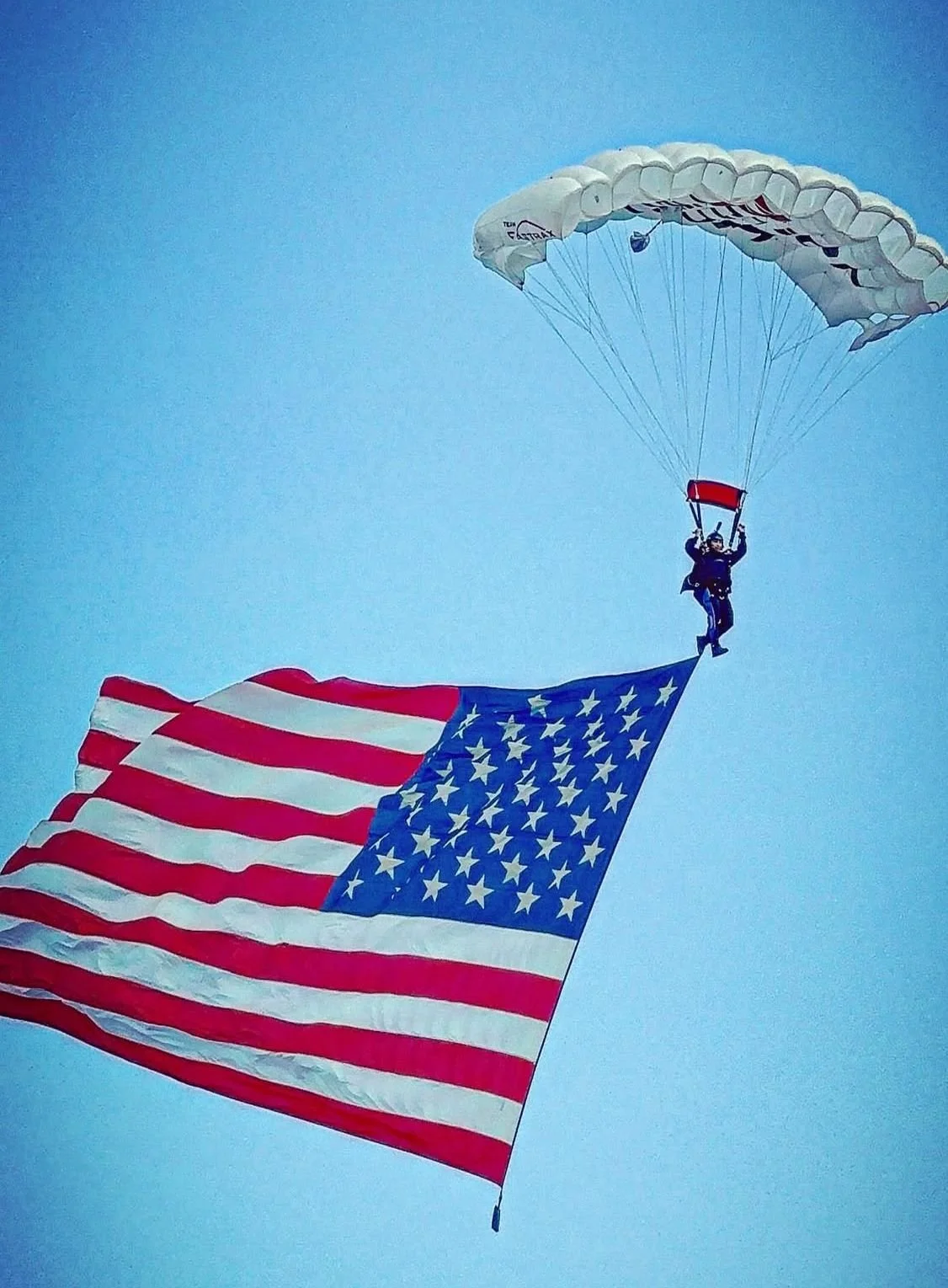 A parachutist descending with a large American flag attached to their parachute against a clear blue sky.