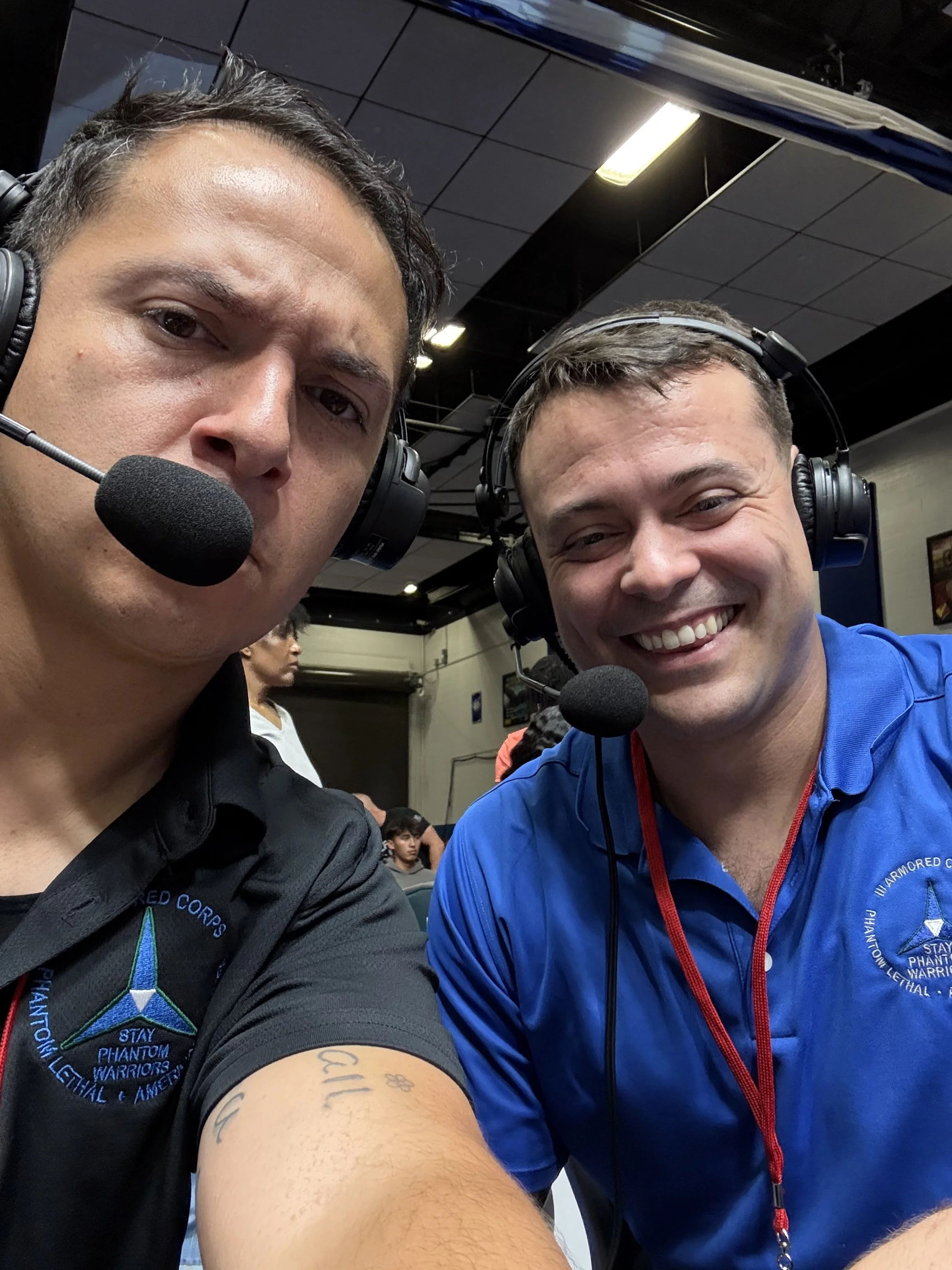 Two men wearing headsets and microphone, smiling and taking a selfie at an indoor event with a dark ceiling and other attendees in the background.