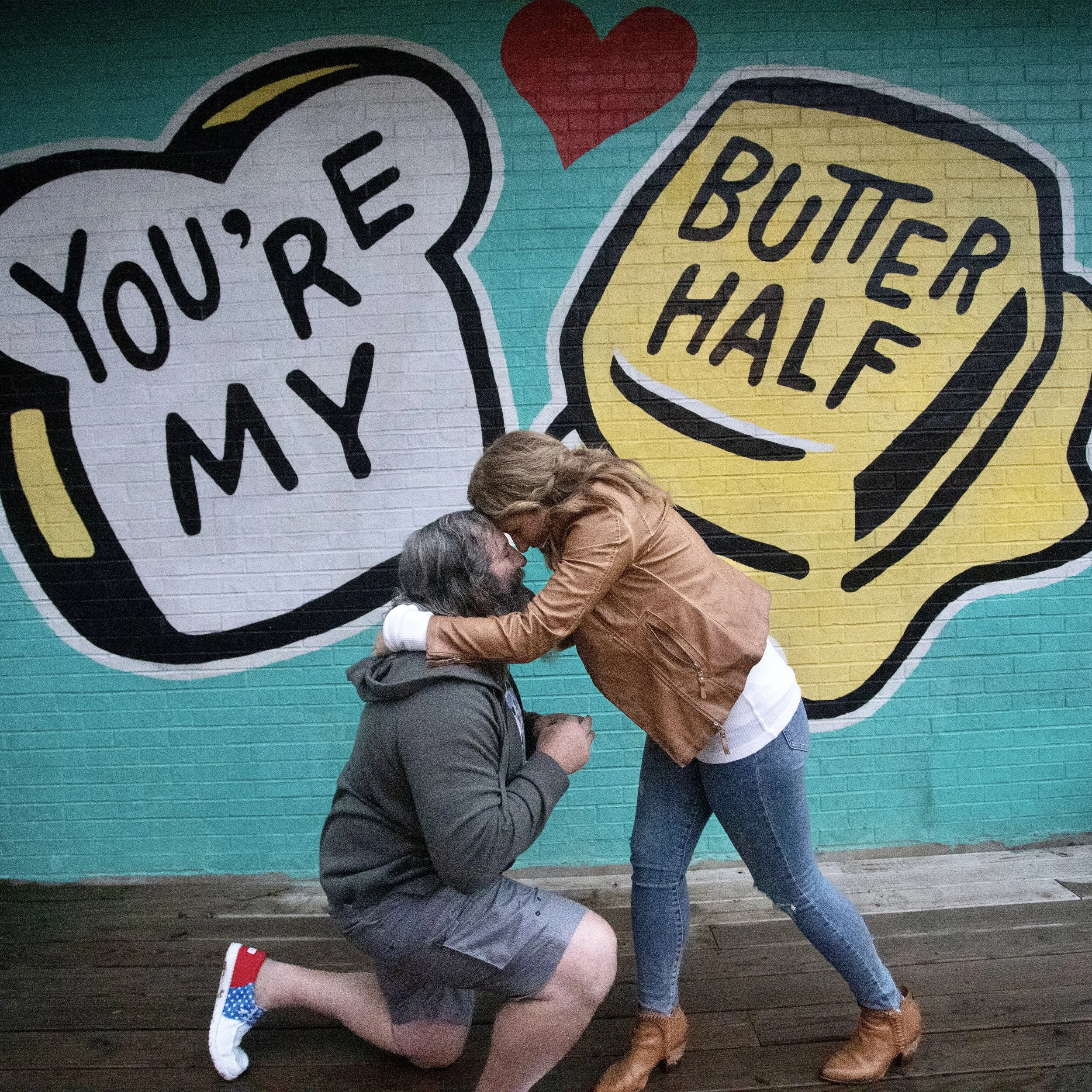 Two people are in an embrace in front of a colorful street mural with speech bubbles and a red heart. One person is squatting, and the other is leaning over them, touching their foreheads. The patterned clothing and background suggest a casual, expre