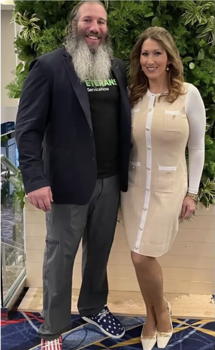 A man with a beard and long hair, wearing a black T-shirt and a blazer, stands next to a woman with long wavy hair, wearing a cream-colored button-up dress. They are smiling and standing indoors in front of green foliage.