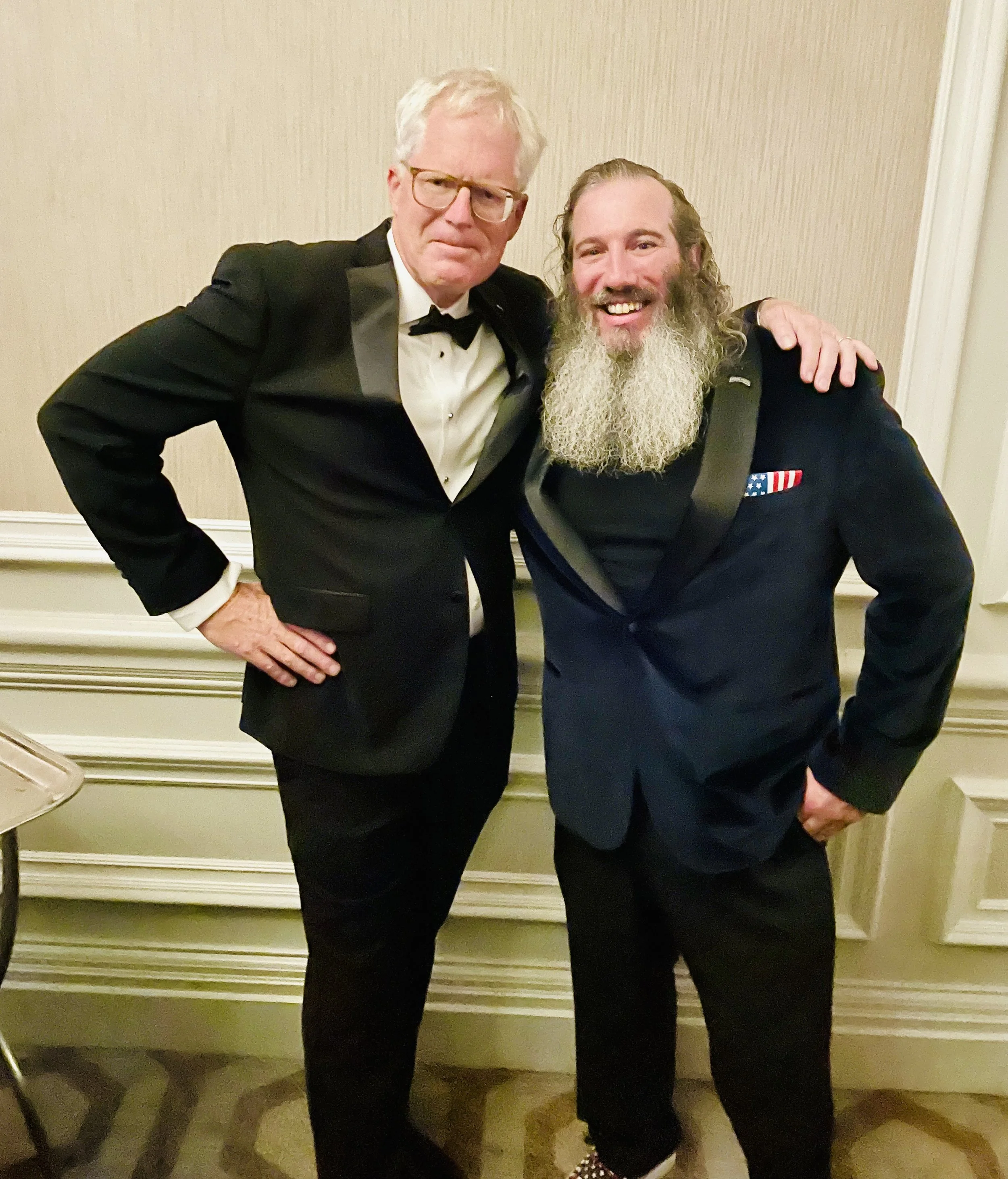 Two men pose for a photo at a formal event, dressed in tuxedos, with one man having white hair and glasses, and the other with long curly hair and a beard, standing in front of a beige patterned wall.