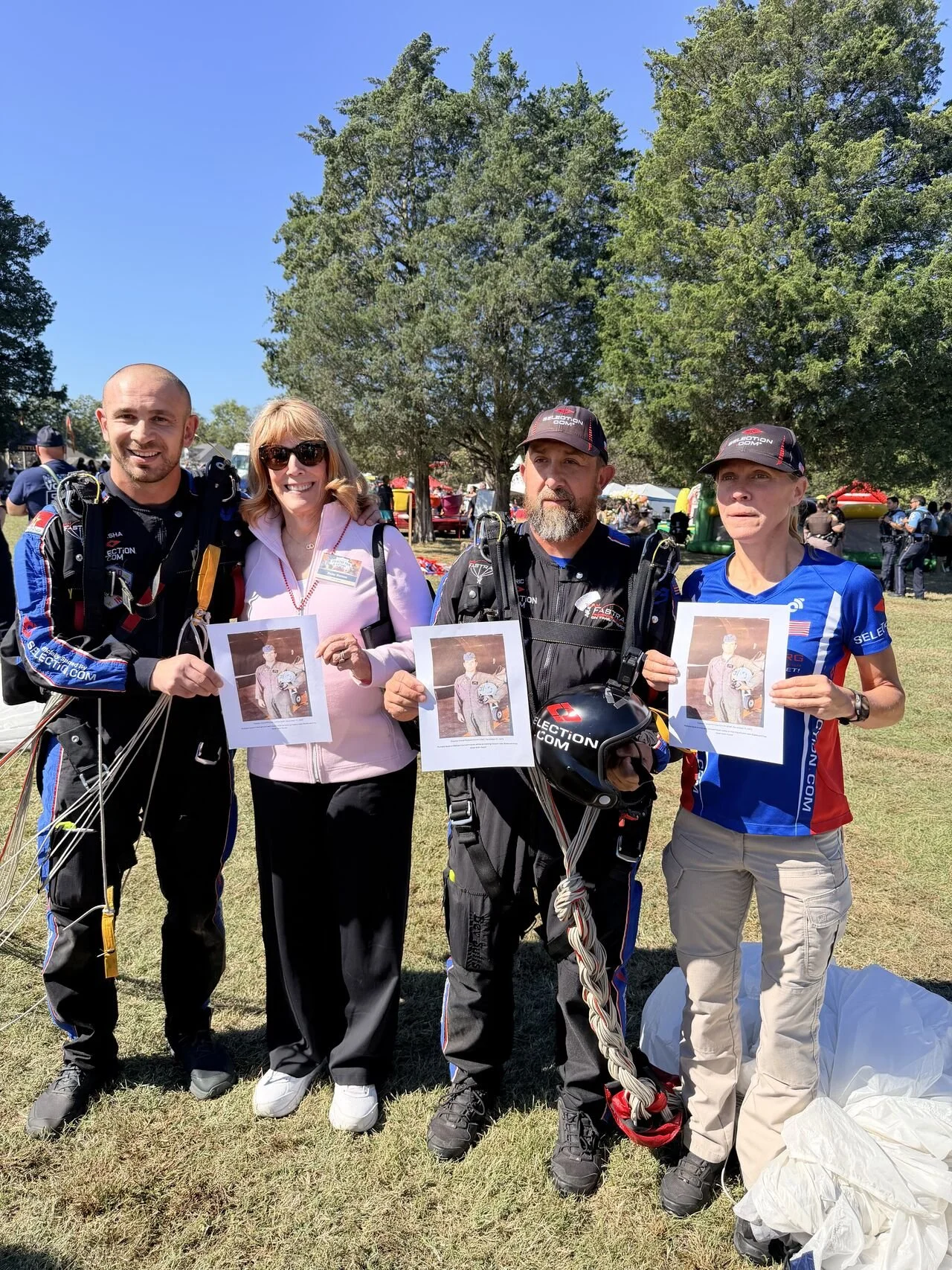 Four people standing outdoors on grass, holding printed photographs of a person in a racing suit. The group appears to be at a racing event, with tents and other people in the background. Two of the individuals are dressed in racing gear, while the o