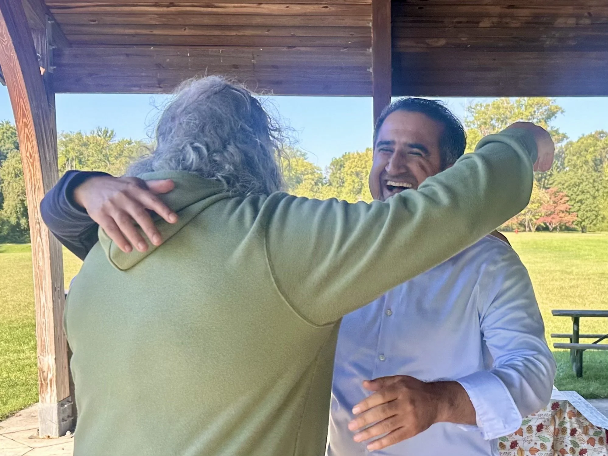 Two people are hugging outdoors under a wooden pavilion, with one person facing away from the camera and the other smiling broadly. The background features trees and a sunny sky.