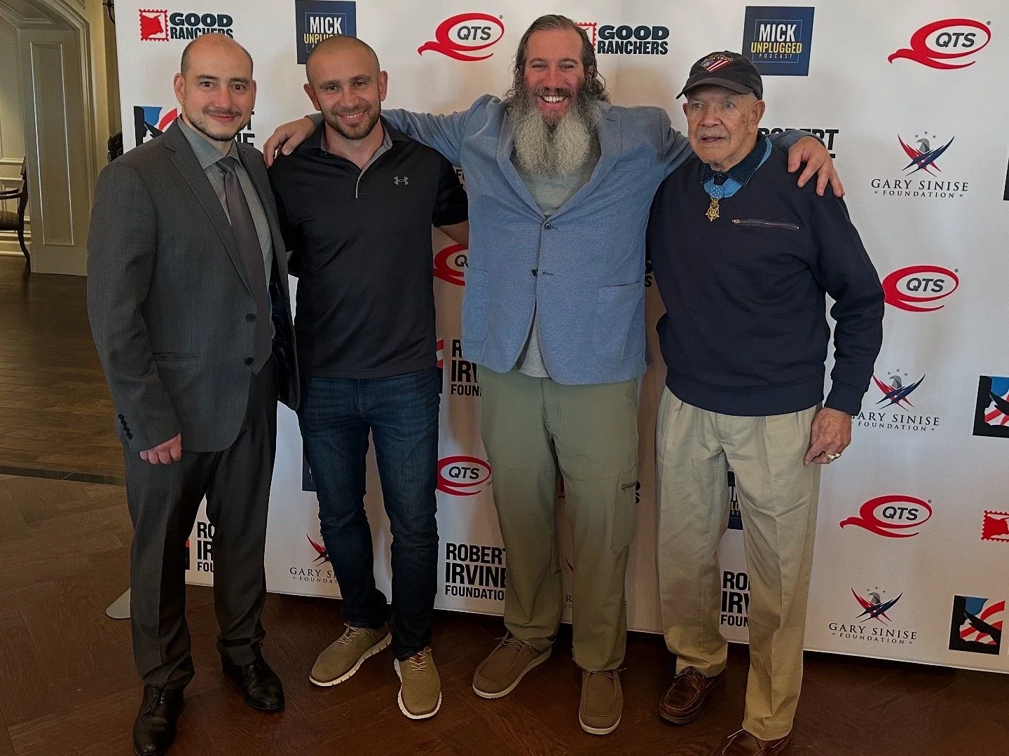 Five men standing together and smiling at an event with a backdrop that has logos for the Gary Sinise Foundation, QTS, and Robert Irvine Foundation. They are dressed in formal and casual attire.