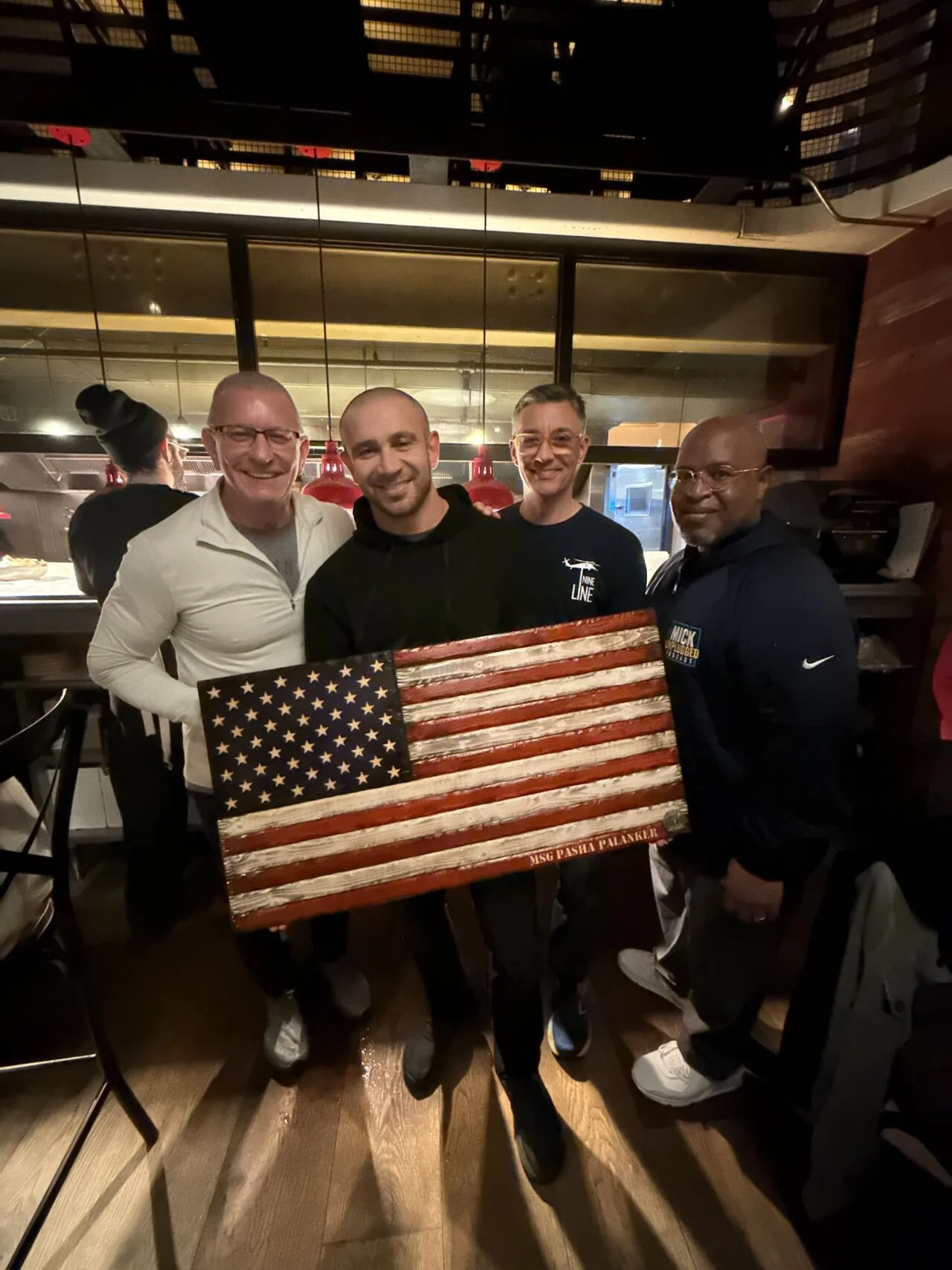Four men standing in a restaurant holding a painted American flag with a message "MISC Pasha Palankai" at the bottom. They are smiling and posing for the photo.