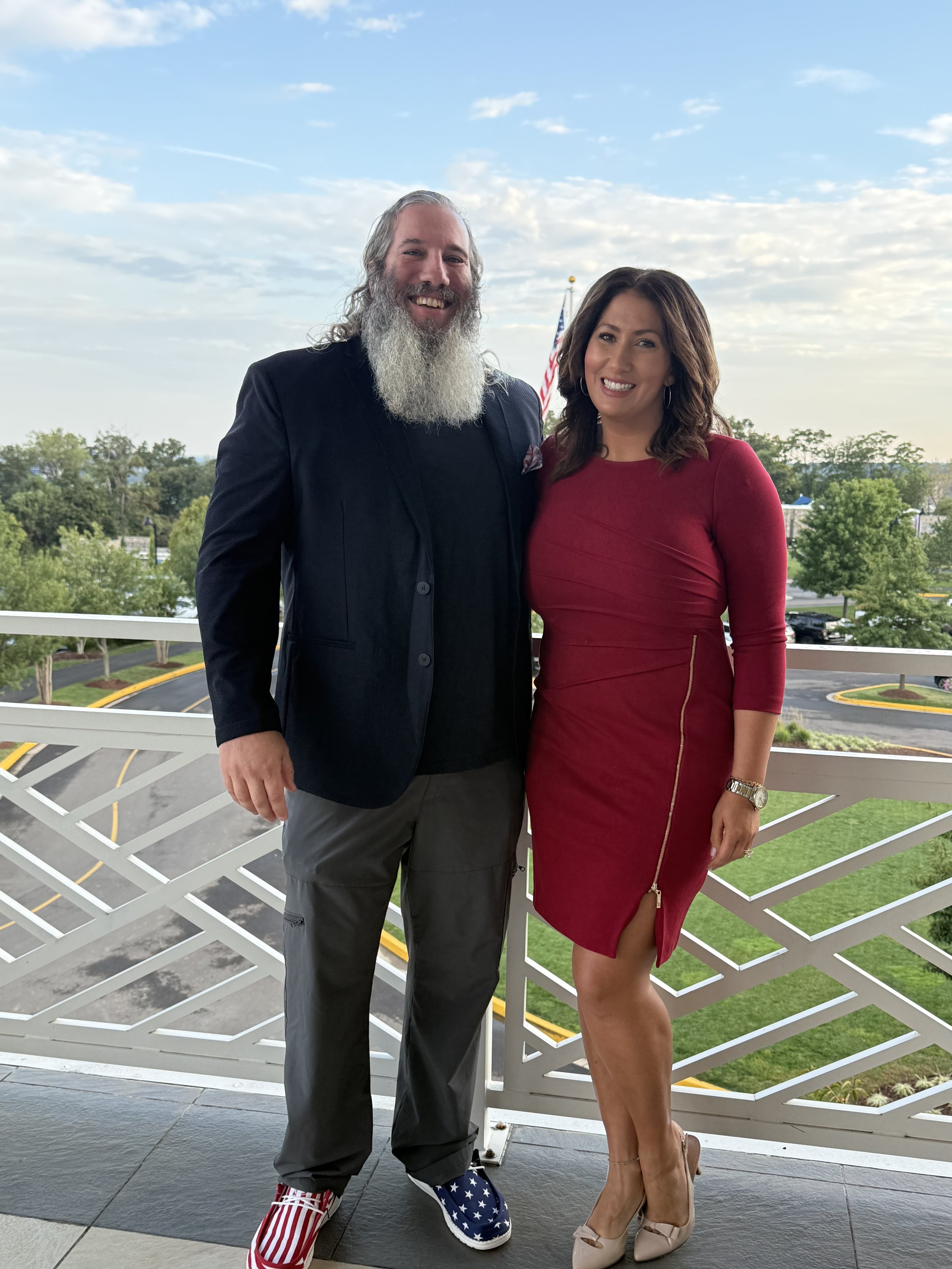 A man with a gray beard and a woman with shoulder-length brown hair standing together on a balcony with a view of greenery and parking lot, both smiling, dressed in semi-formal attire, with the man wearing patriotic shoes and the woman in a red dress