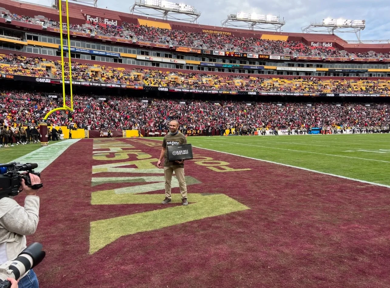 A man standing on a football field with a large crowd in the stands behind him, holding a black box. A cameraman is filming him, and the stadium has yellow and maroon colors with the logo of the Washington football team.