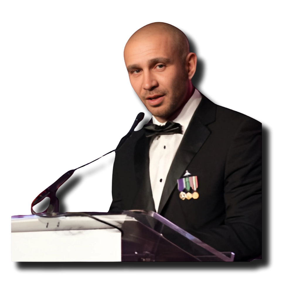 A man in a tuxedo with medals on his chest speaking at a podium with a microphone.