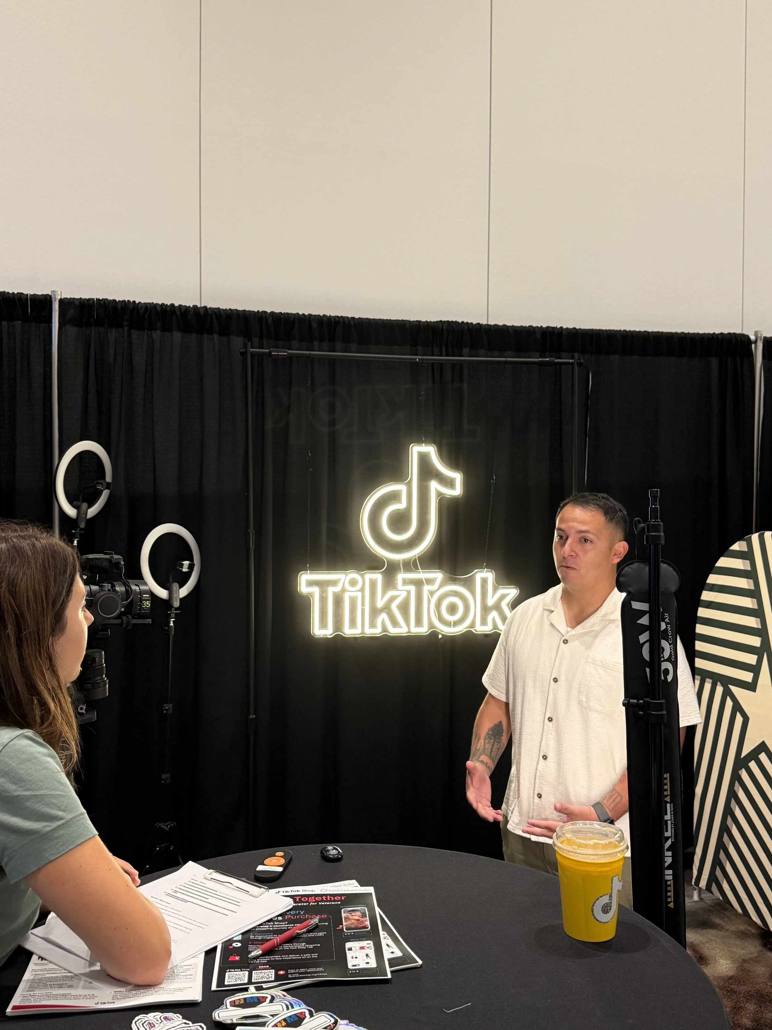 A man in a white shirt is speaking at a TikTok booth, with a black curtain behind him featuring a neon TikTok logo. A woman with brown hair is sitting at a table with brochures, papers, and a red pen. There is a yellow drink with a lid and a straw on the table. The setting appears to be a conference or expo.