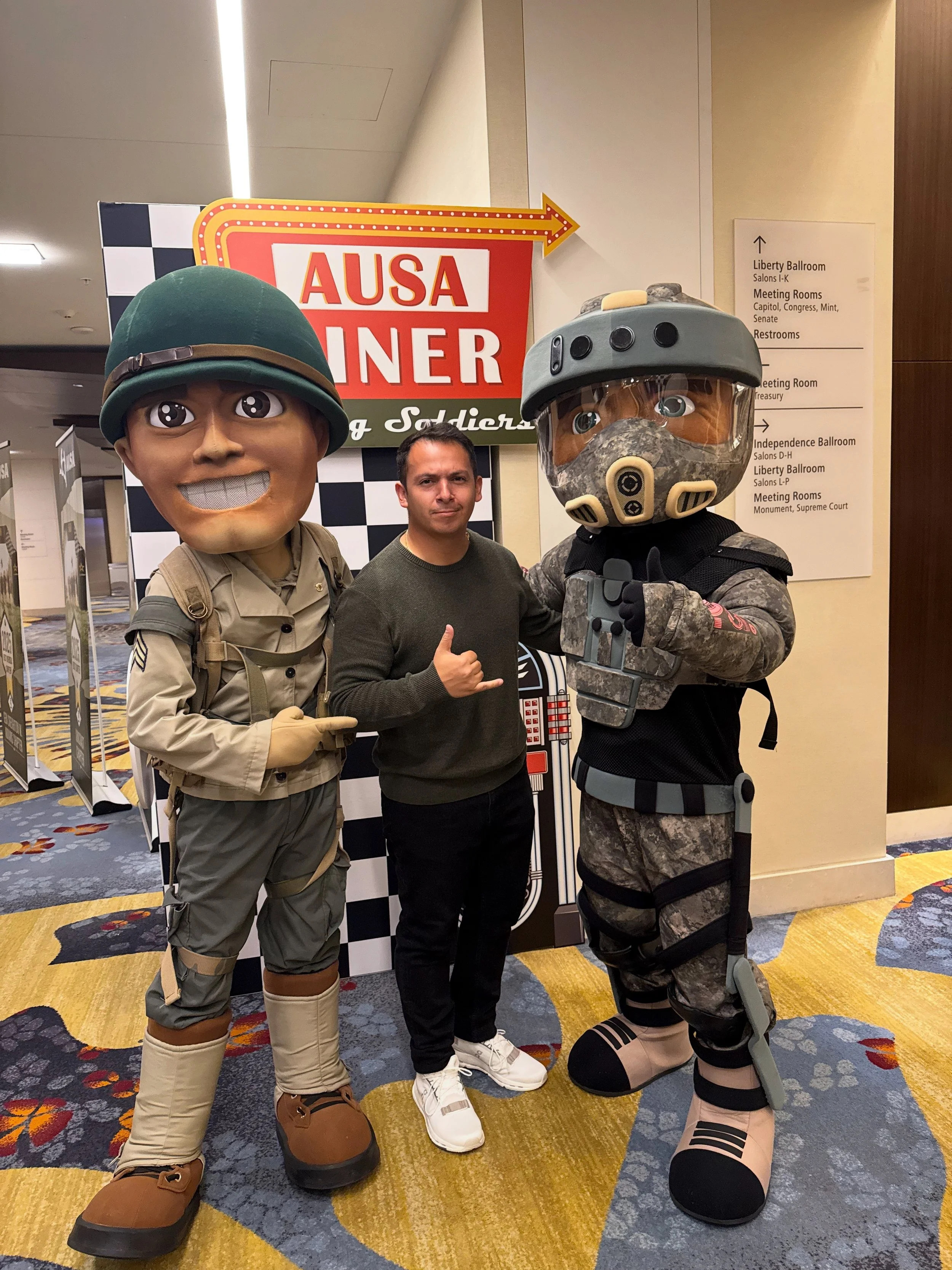 A man standing between two people in military costumes at an indoor event. The costumes include oversized helmets, military uniforms, and large boots. The background has a sign with the text 'AUSA' and a checkered pattern.