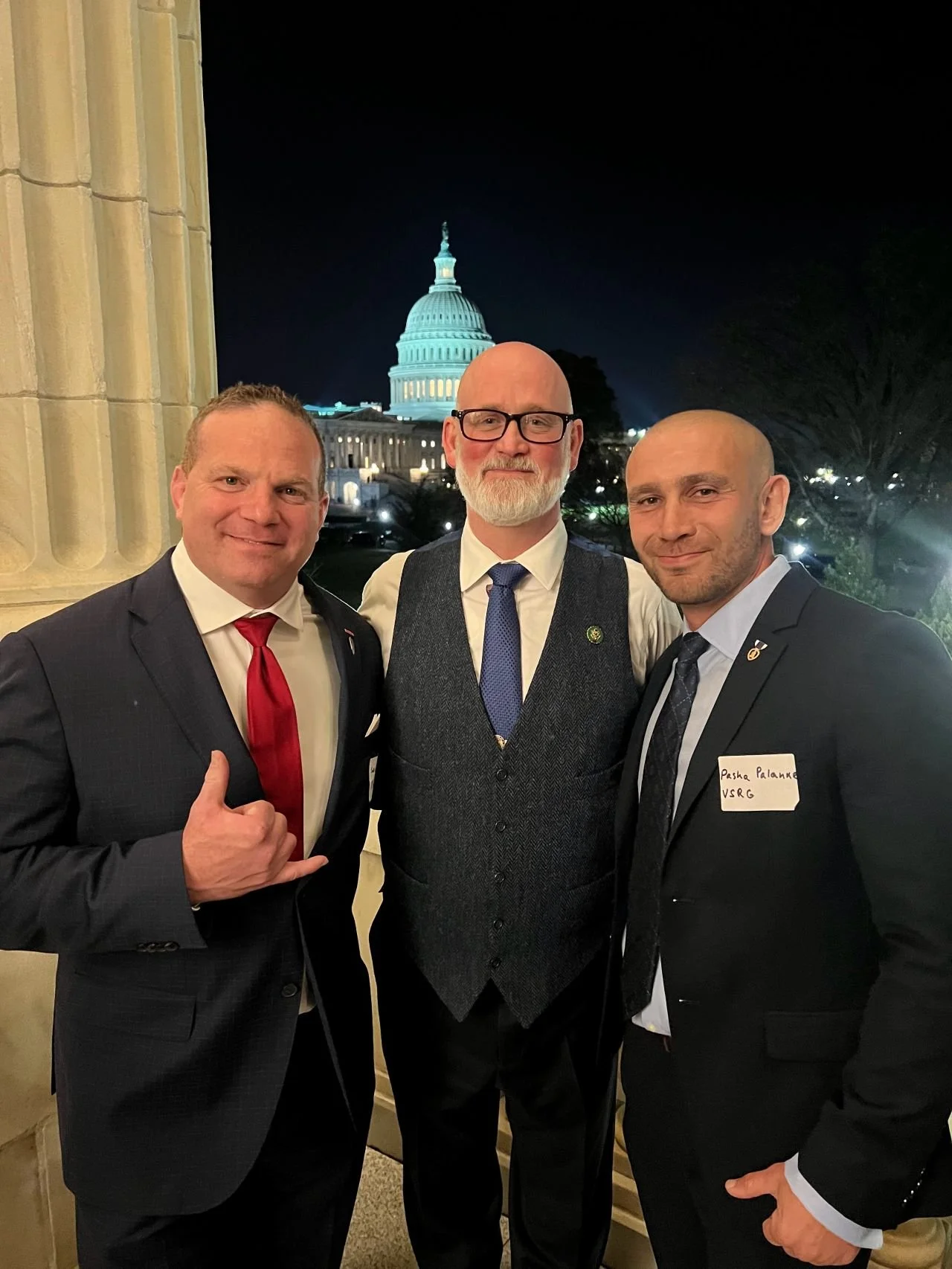 Three men in suits standing together at night with the U.S. Capitol building illuminated in the background. The man on the left is giving a thumbs-up gesture. The man in the middle is wearing glasses, a white shirt, a blue tie, and a gray vest. The m
