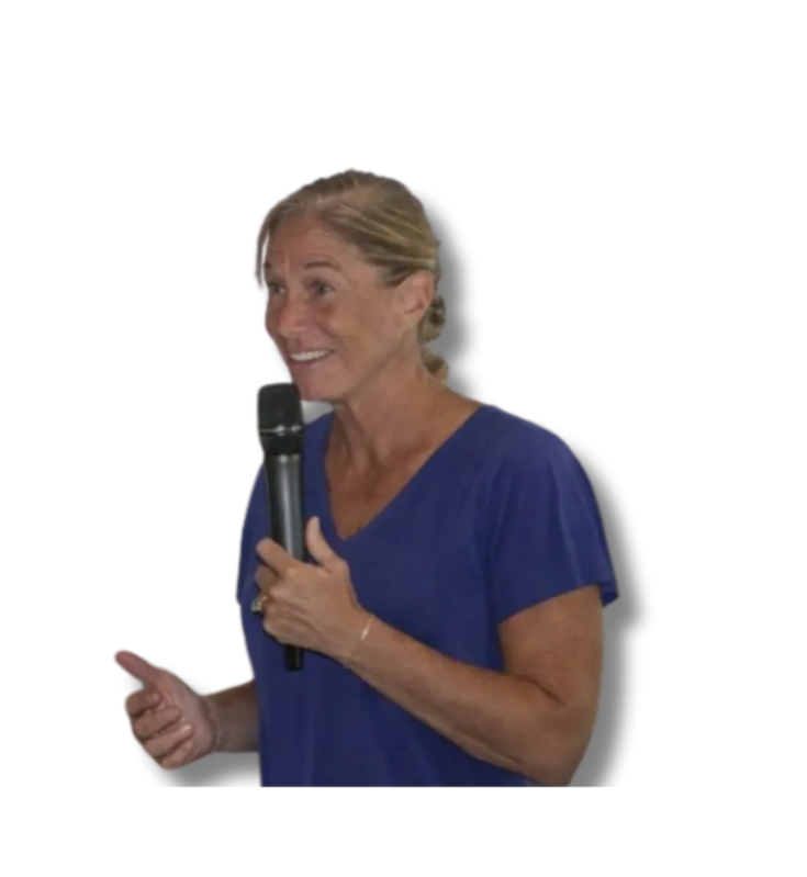 A woman with blonde hair in a ponytail, wearing a blue shirt, holding a microphone, smiling, and speaking.