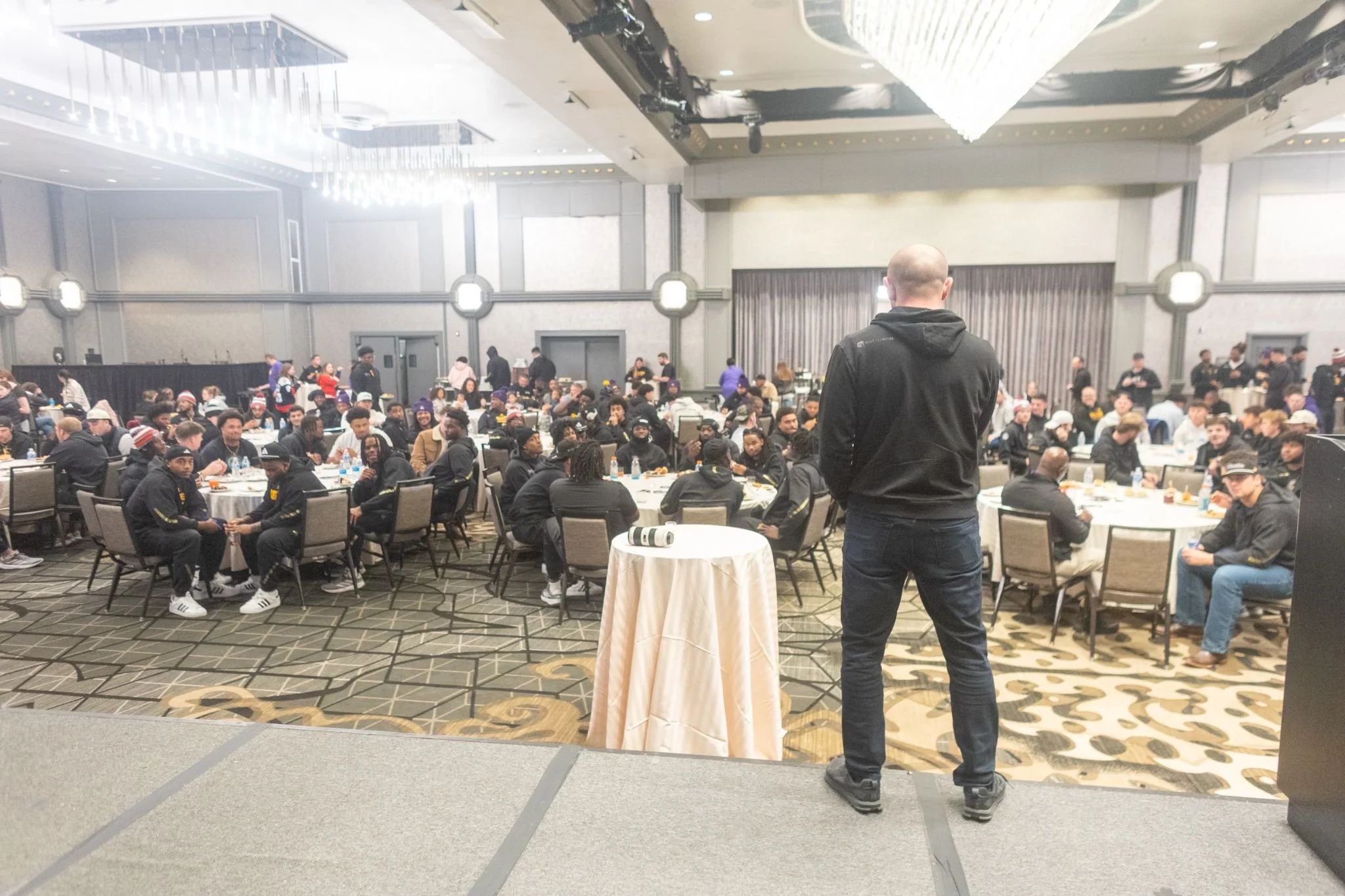A large conference or banquet room filled with many seated attendees. A man with a shaved head, dressed in black, stands facing the audience from a small table at the front. The room has high ceilings, chandeliers, and a patterned carpet.