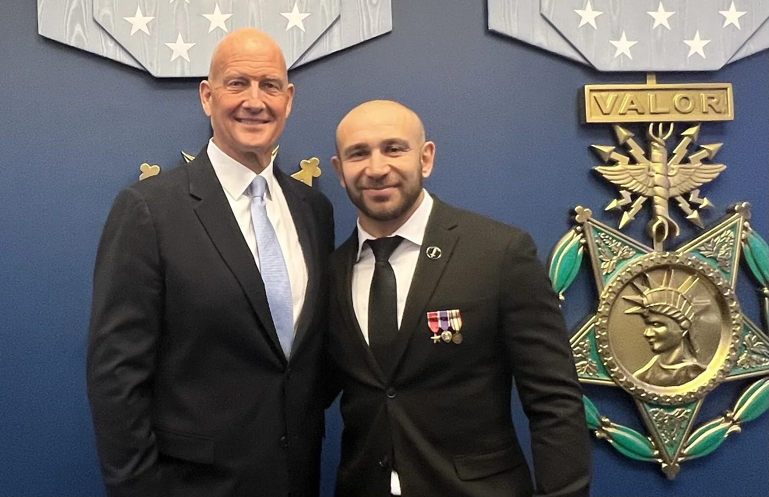 Two men in formal suits standing in front of military-inspired wall decor with a large medal and the word 'VALOR' on a plaque. One man is taller, bald, and smiling, while the shorter man has a beard and medals on his suit.