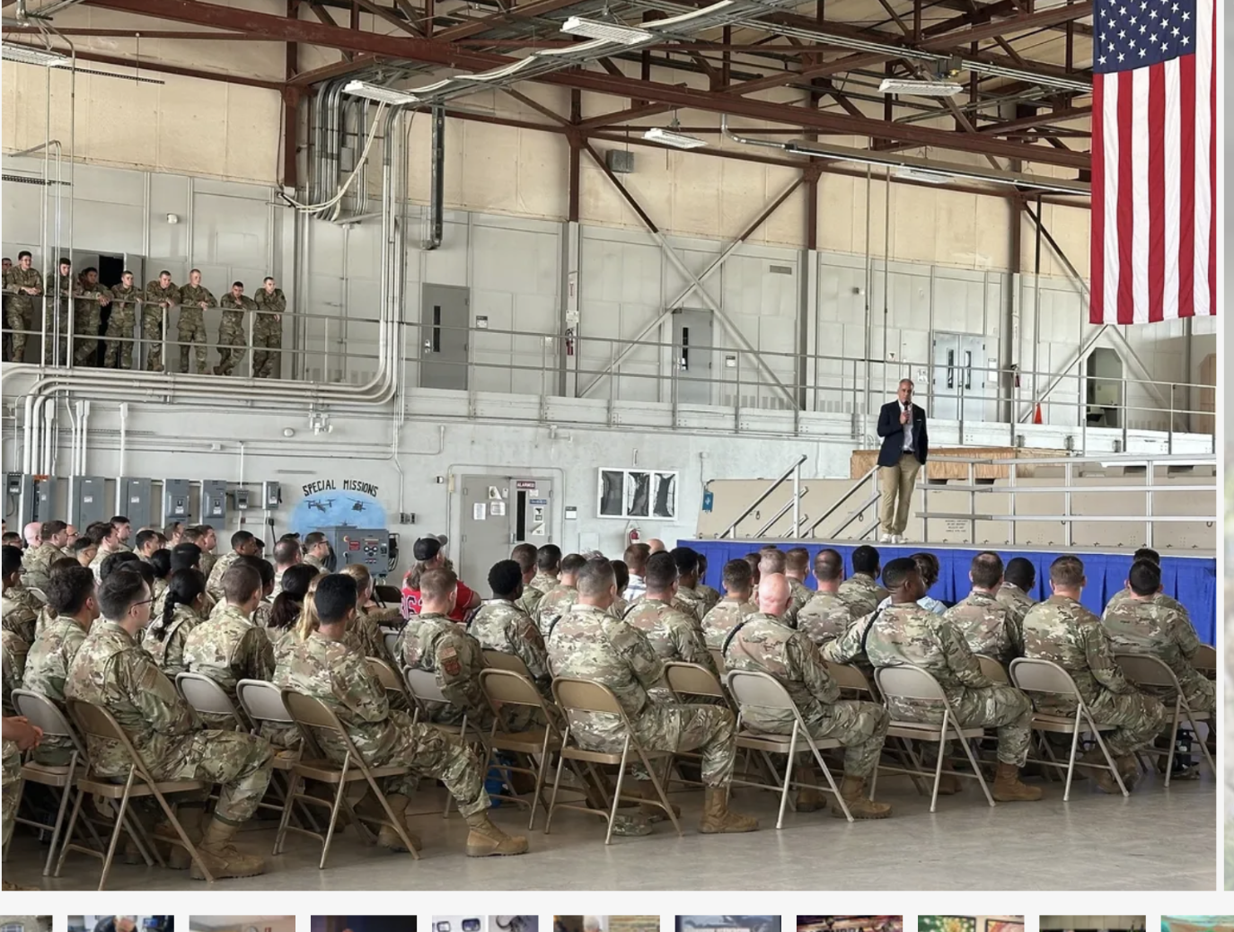 Military personnel seated in an indoor hangar listening to a speaker at a podium. There are soldiers on a balcony behind, and an American flag hanging from the ceiling.