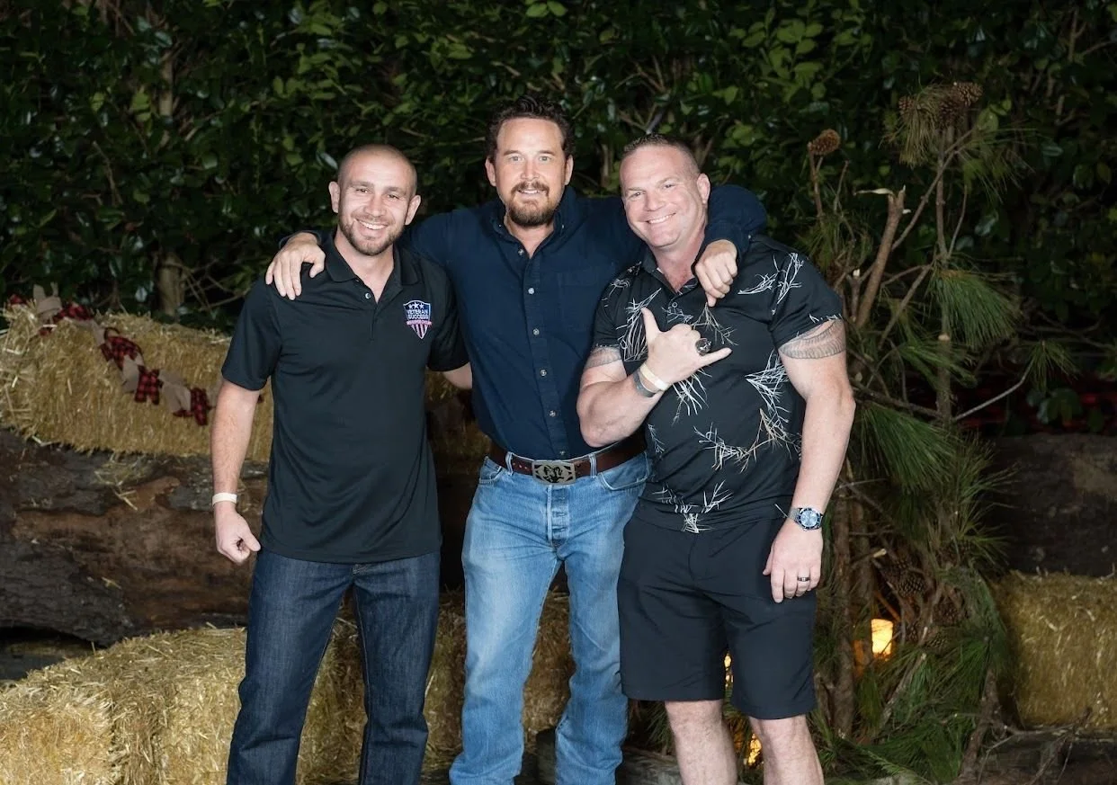 Three men standing outside at night, smiling, with greenery and hay bales behind them. One man on the right is making a hand gesture.