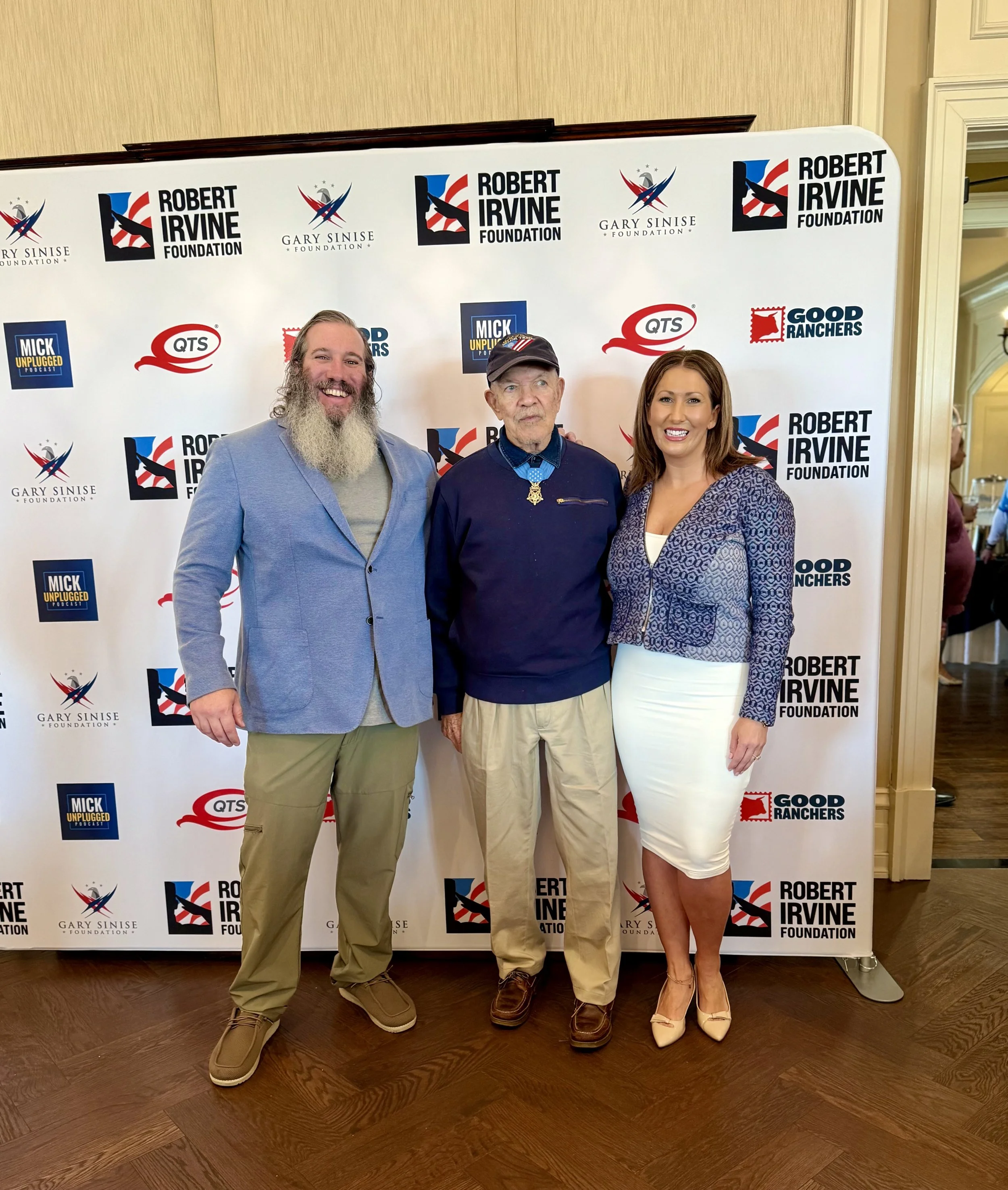 Three people standing in front of a promotional backdrop with logos, smiling at the camera. The man on the left has long hair and a beard, wearing a light blue blazer and khaki pants. The man in the middle has short hair, wearing a dark jacket and a 