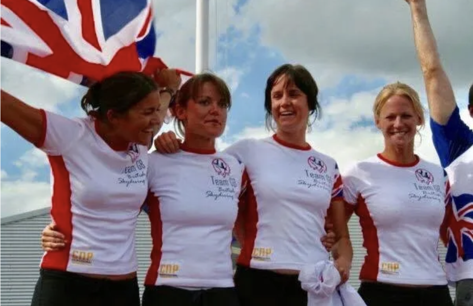 Female athletes in white shirts with red accents celebrate together, some holding a British flag, outdoors under a partly cloudy sky.
