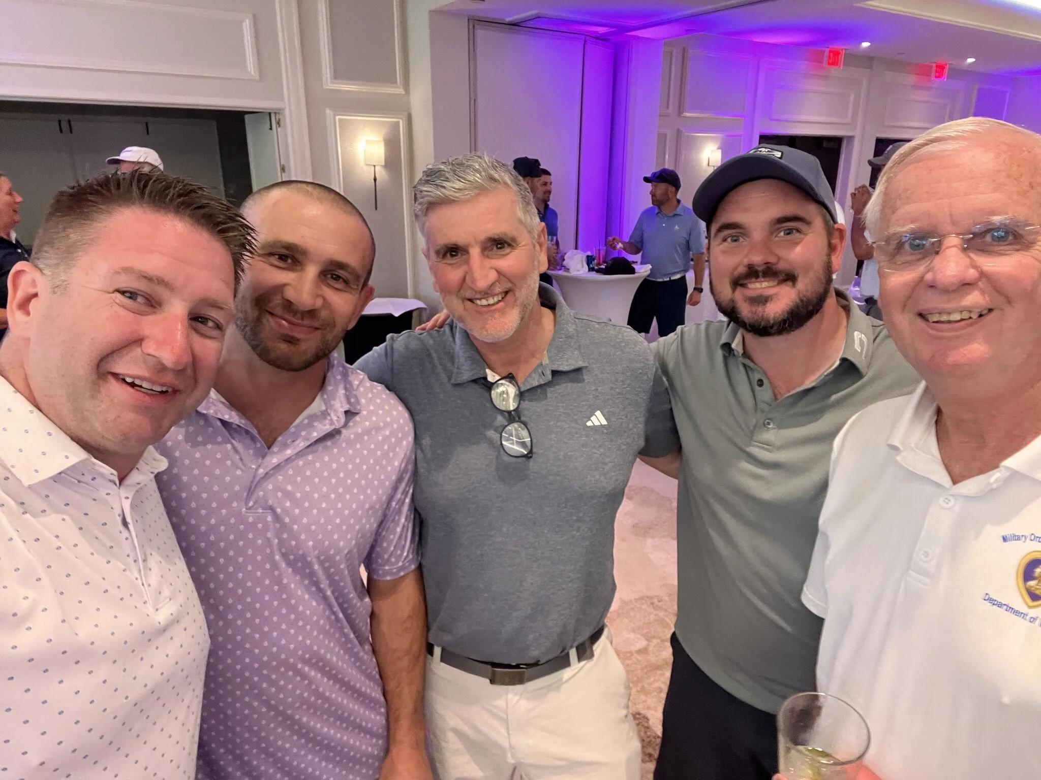 Group of five men smiling at a social event, standing close together in a banquet hall with purple lighting, with other guests in the background.